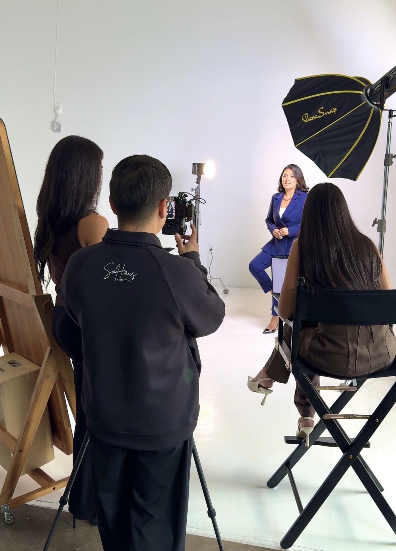 Person filming woman in blue suit during interview in studio with lighting equipment, white background, and chairs.