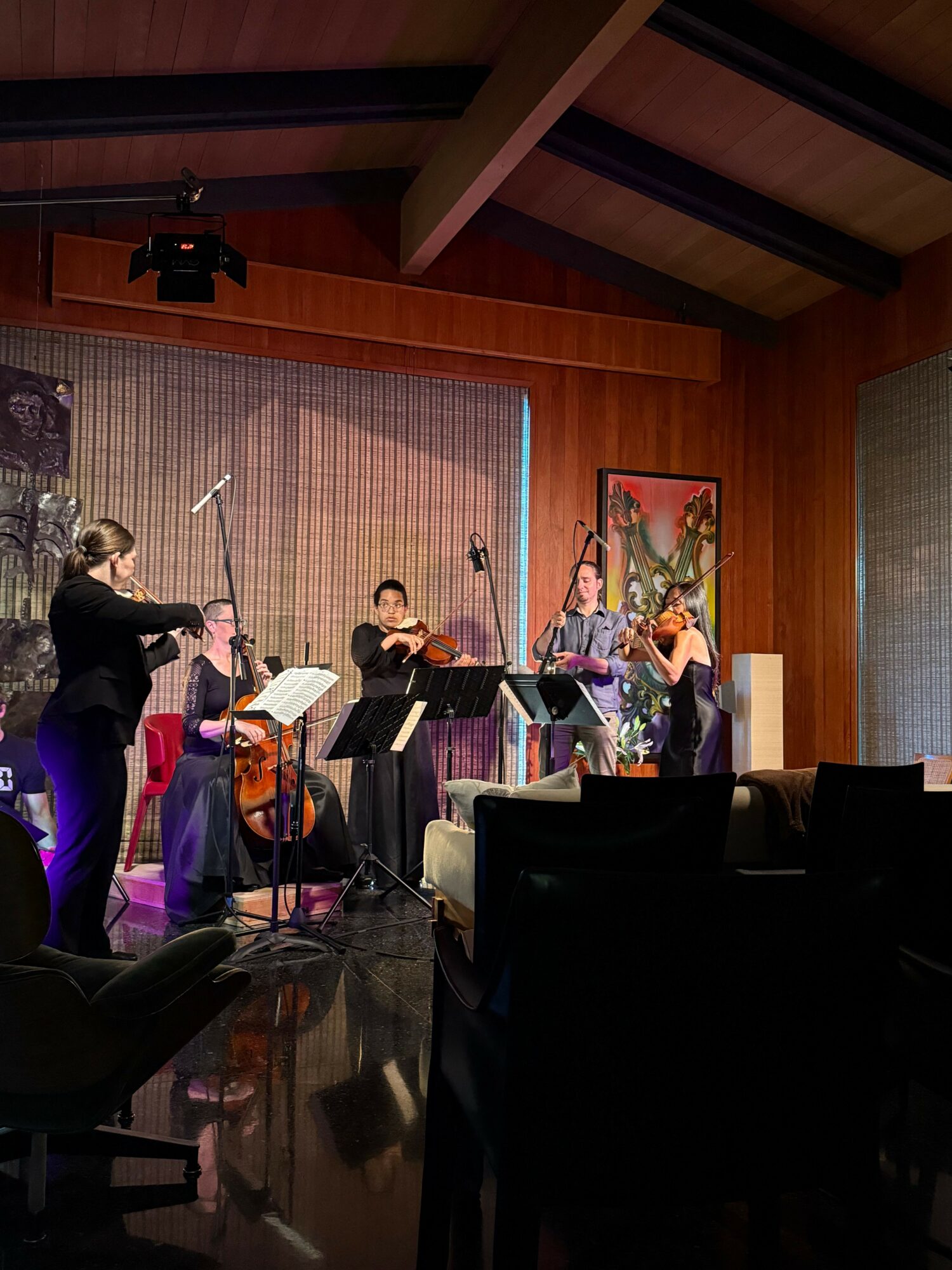 People gathered around a table in a room with wooden walls and a colorful painting, some holding papers or instruments.