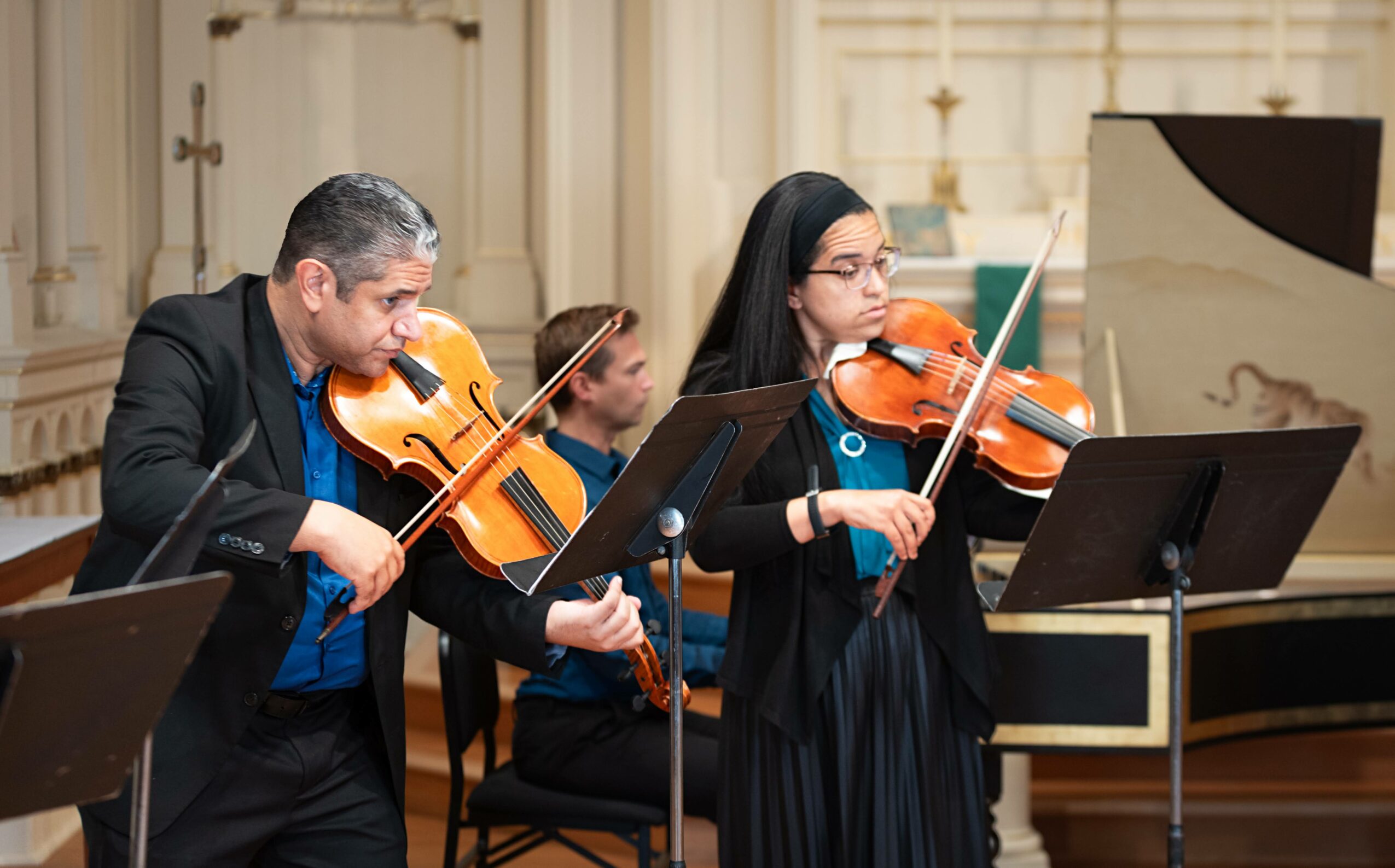 Two musicians playing violins in a concert hall, with music stands and a piano in the background.
