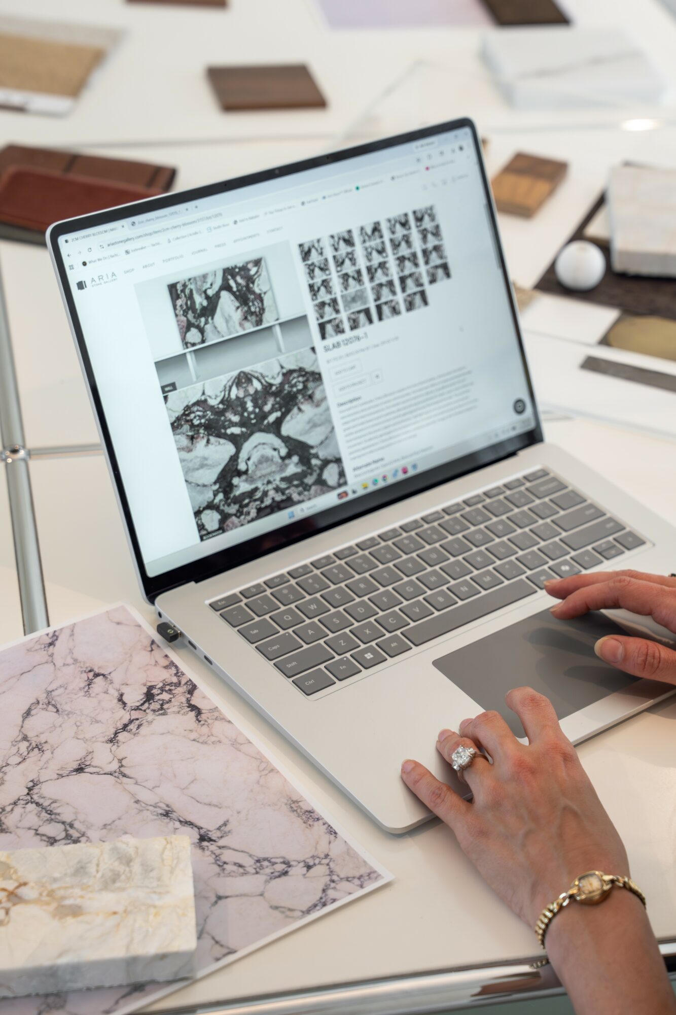 Person using a laptop with images and text on the screen, on a white desk with marble-patterned paper and wooden samples.