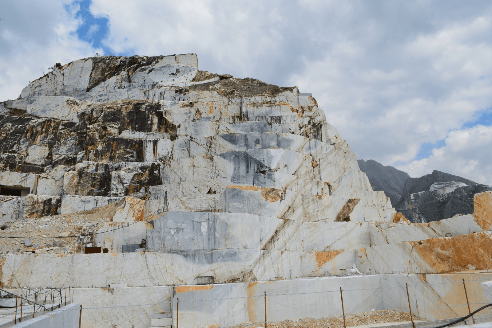Marble quarry with large white and gray stone blocks, mountain landscape in background, partly cloudy sky.