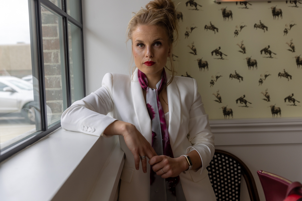 Woman with blonde hair in a white blazer sitting by a window, wallpaper with animal silhouettes in background.