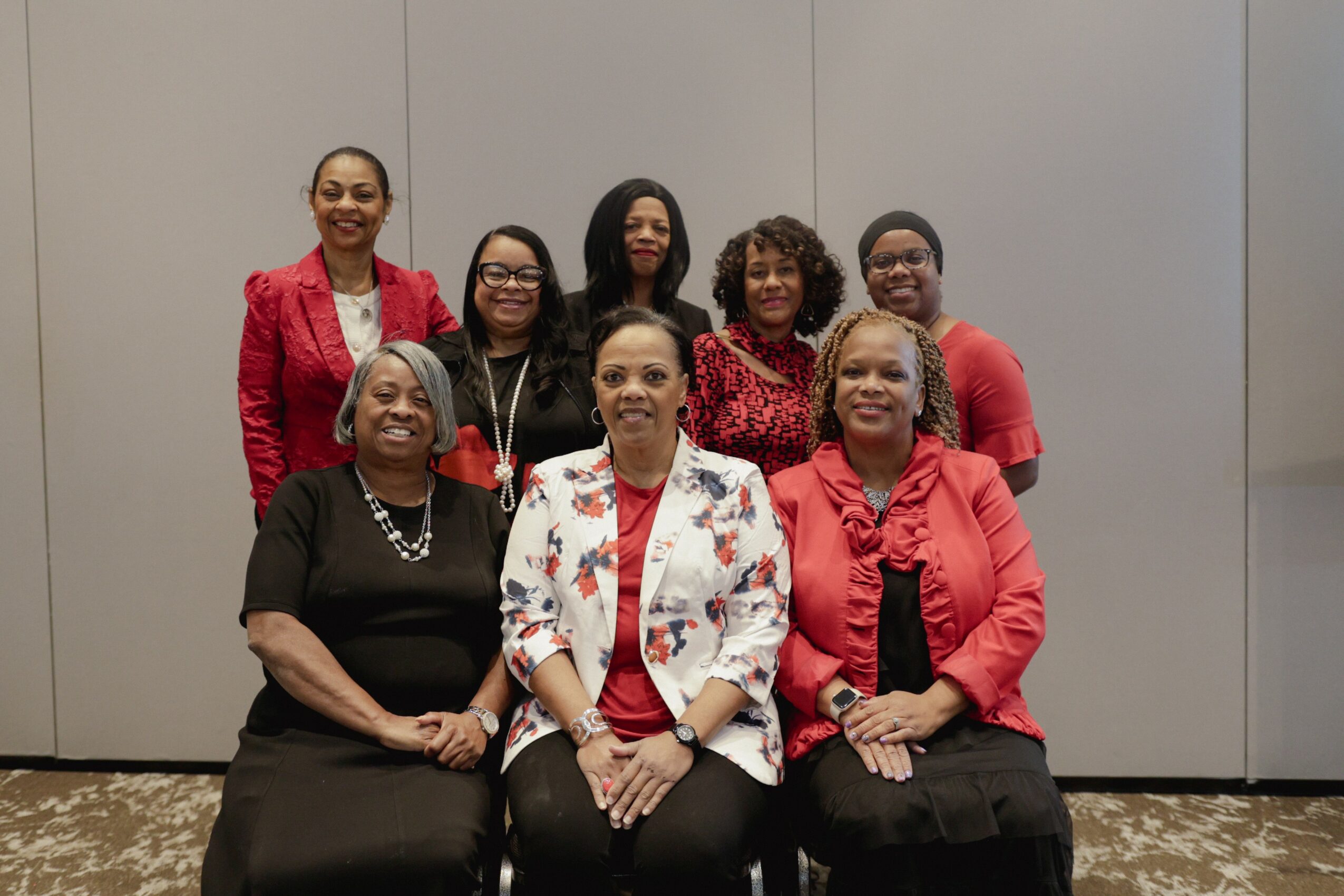 Group of eight women standing and sitting in front of a plain wall, smiling, dressed in red, black, and white clothing.