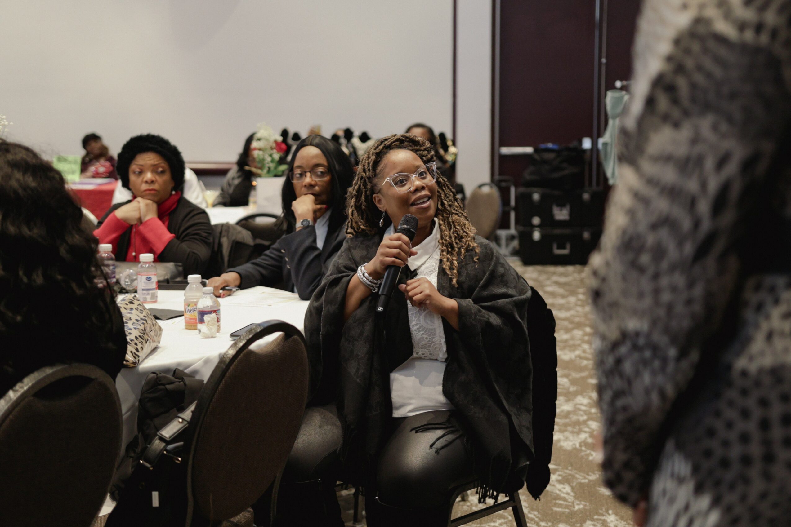 Woman speaking into microphone at a conference table with seated audience members, some taking notes, in a room with a large screen.