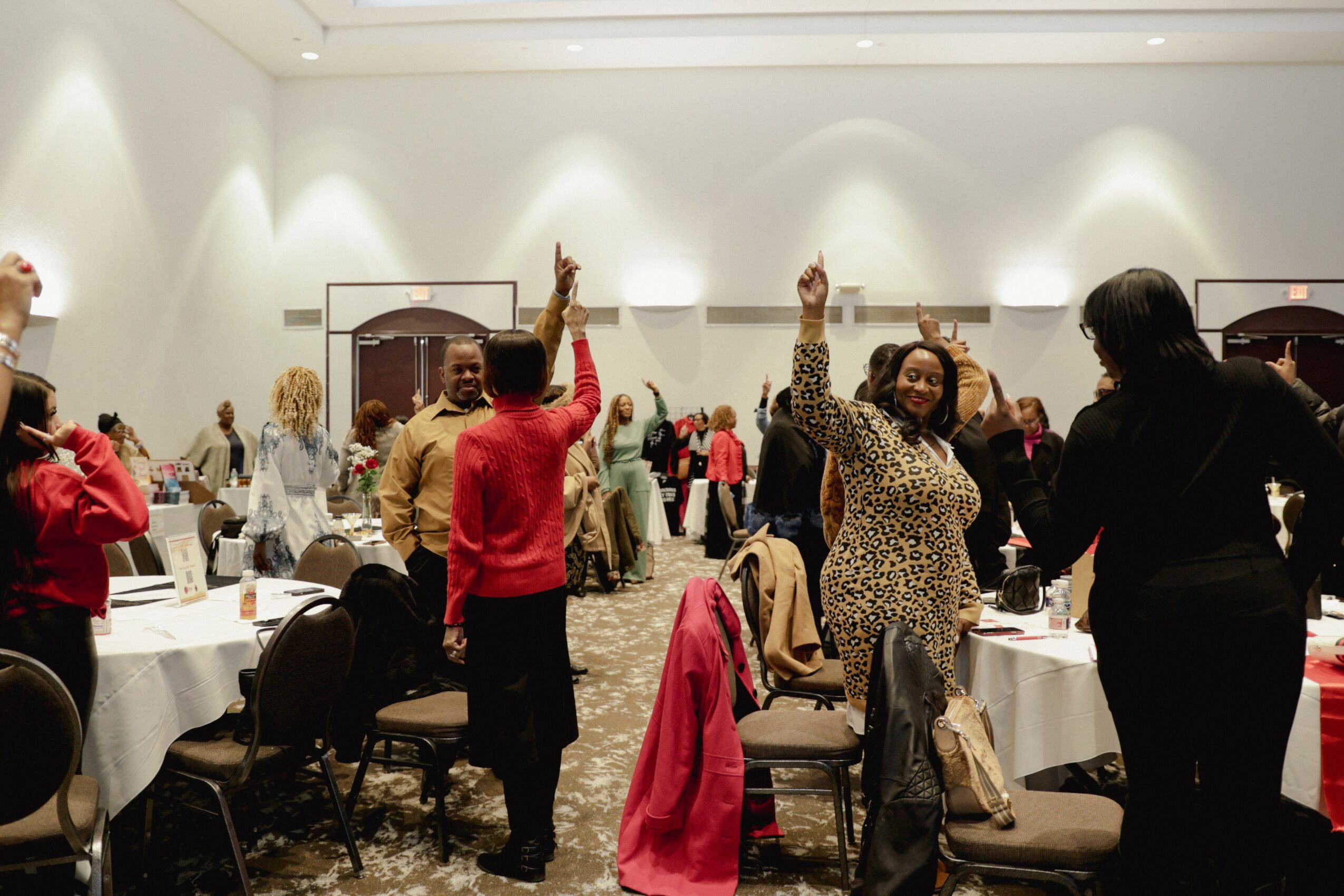 People standing and raising hands in a large, well-lit banquet hall with round tables and chairs, some with pink jackets draped over chairs.