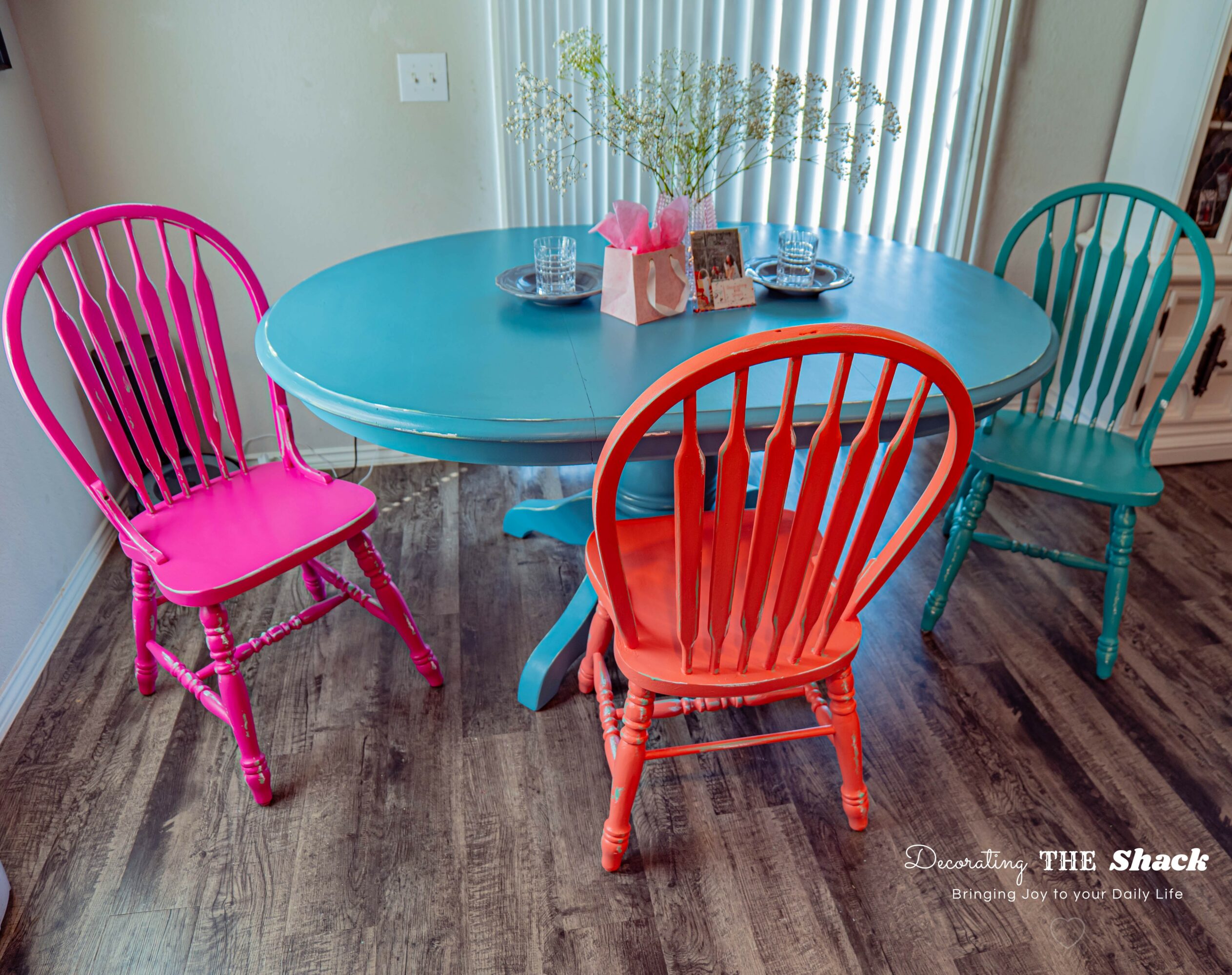Colorful dining table with four chairs in pink, orange, teal, and blue, on a wooden floor near a window with vertical blinds.