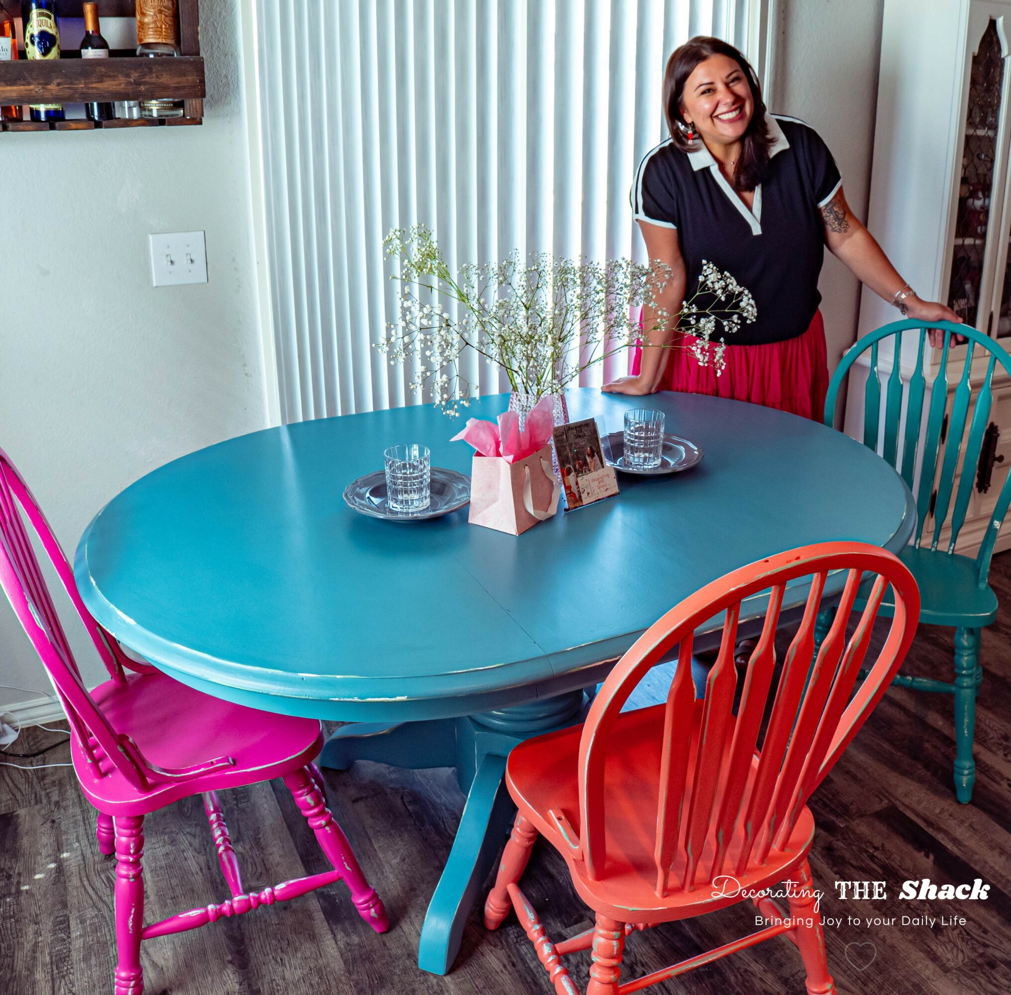 Woman smiling standing next to a blue dining table with colorful chairs, flowers, and tableware, in a bright room.
