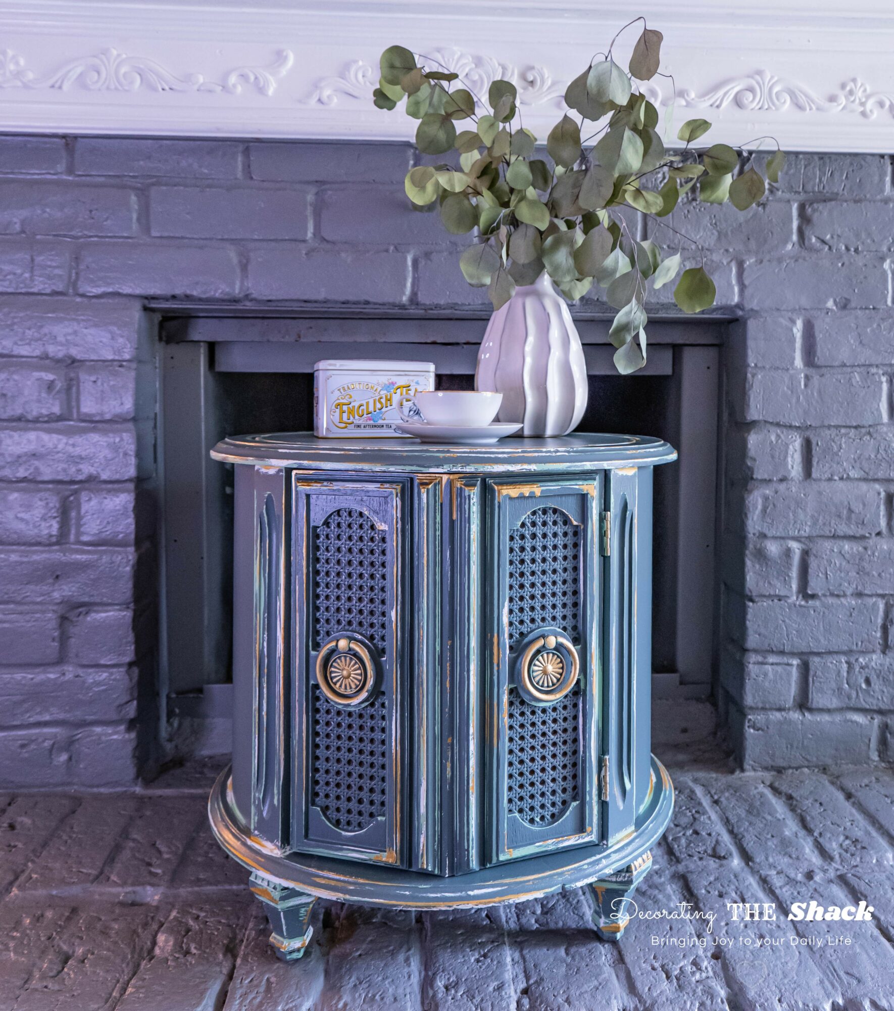 Blue vintage cabinet with a white vase and green leafy plant on top, in front of a brick fireplace, with decorative molding above.