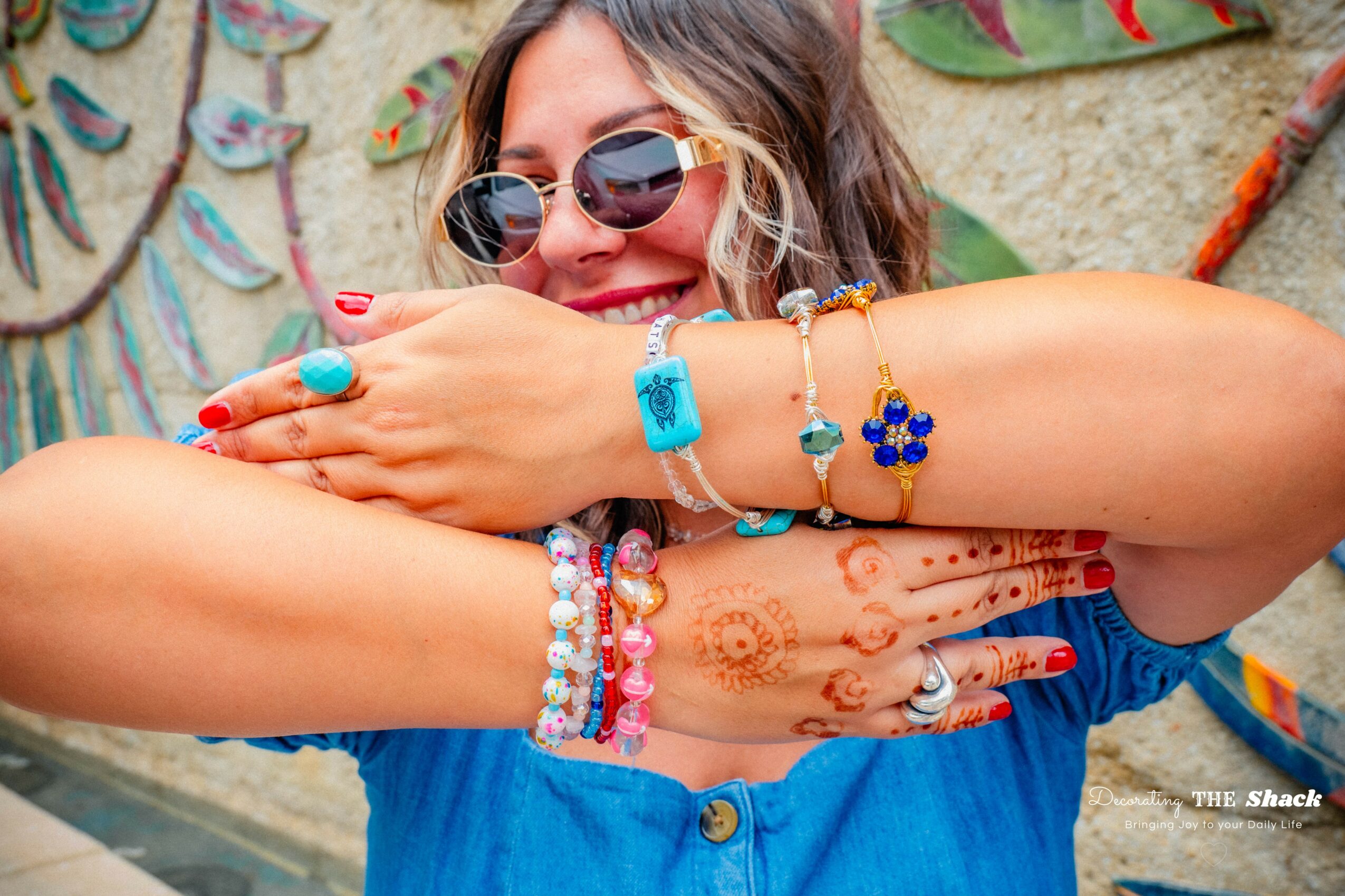Woman with sunglasses smiling, showing colorful jewelry and henna tattoos on her arms, in front of a decorated wall.