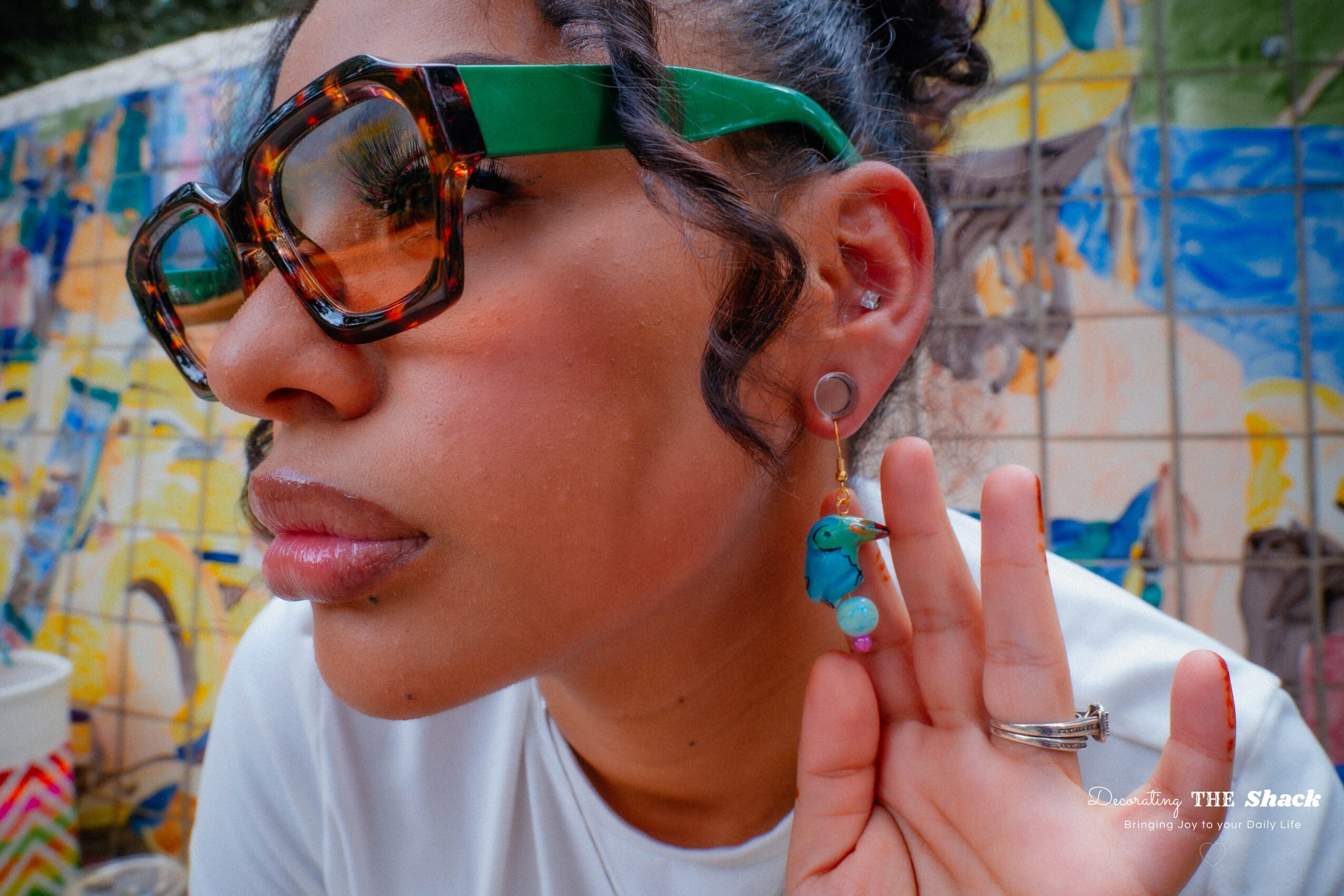 Close-up of a woman wearing large sunglasses, earrings, and a ring, holding her hand near her face against a colorful graffiti background.