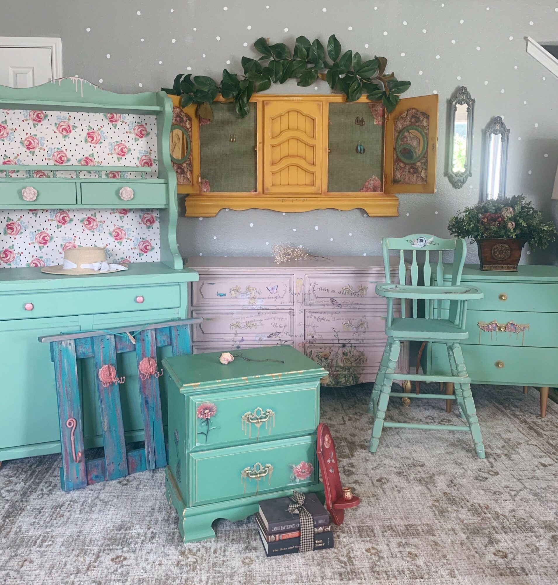 Room with pastel furniture, a wall mirror, and decorative plants, featuring a dresser, chair, and small chest, with a carpeted floor.