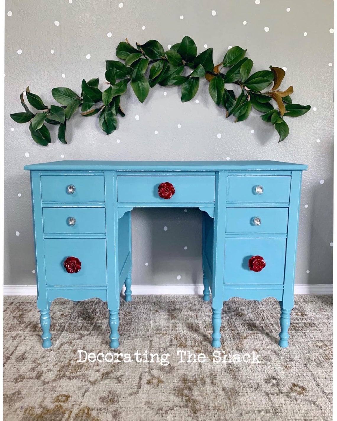Blue wooden desk with six drawers, decorated with red flowers, against a wall with a large green plant above, on a carpeted floor.