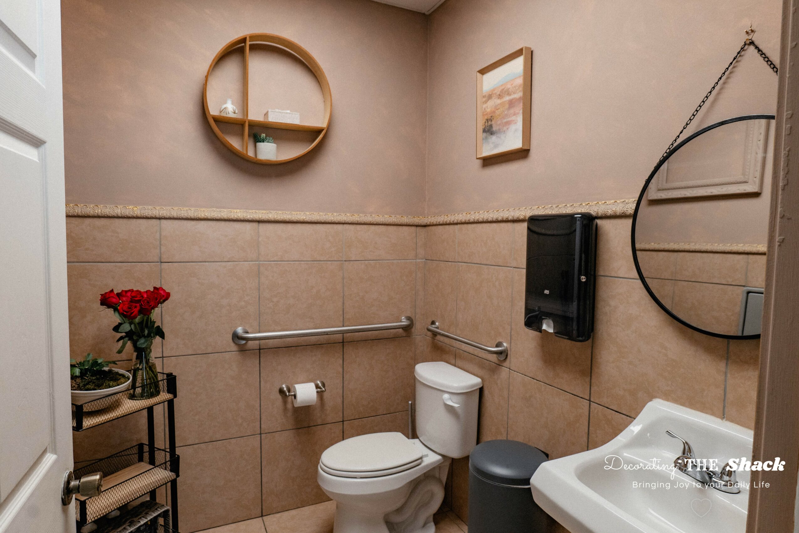 Bathroom with beige tiled walls, toilet, round mirror, wall-mounted black dispenser, and small table with red flowers.