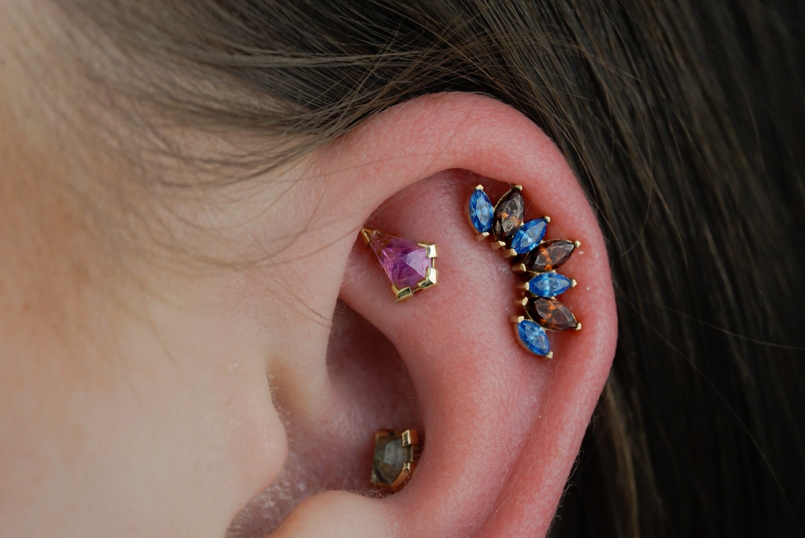 Close-up of a person's ear with multiple colorful earrings, including a pink and purple gem, blue stones, and a greenish gem.