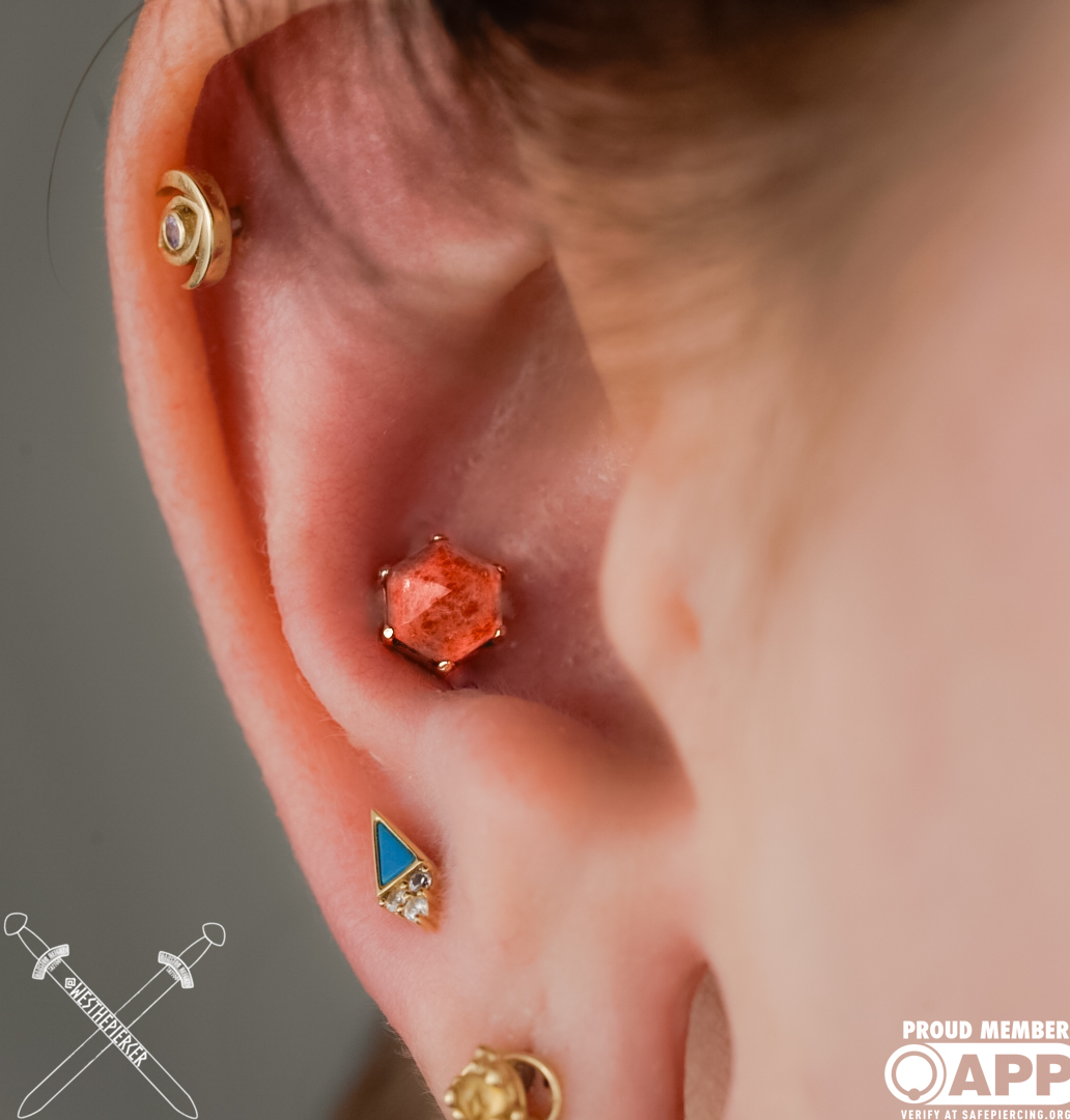 Close-up of a person's ear with three earrings: a gold spiral, a hexagonal orange gemstone, and a triangular blue and gold design.