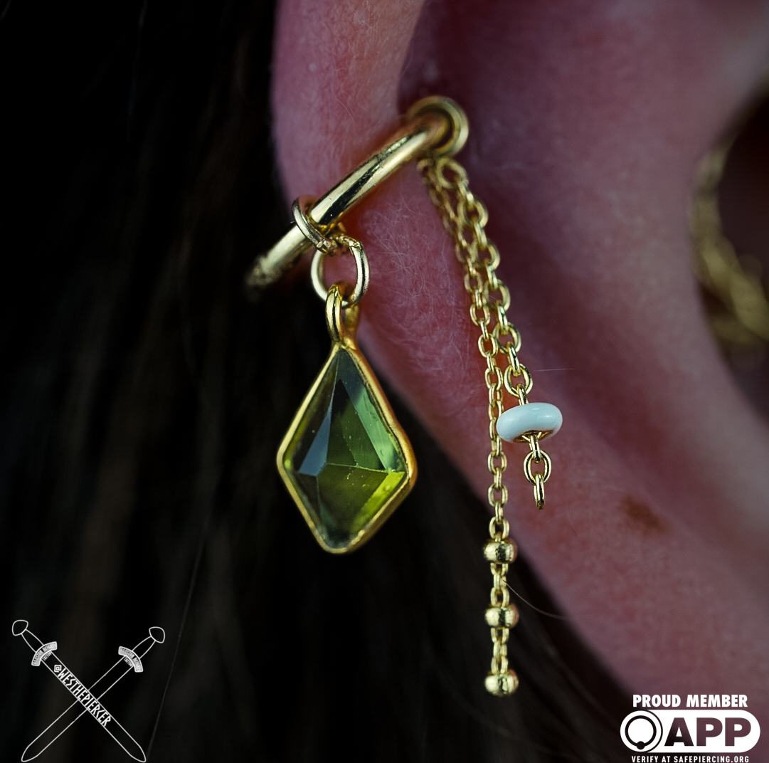 Close-up of a person's ear with multiple gold earrings, including a green gemstone pendant, a white bead, and chains.