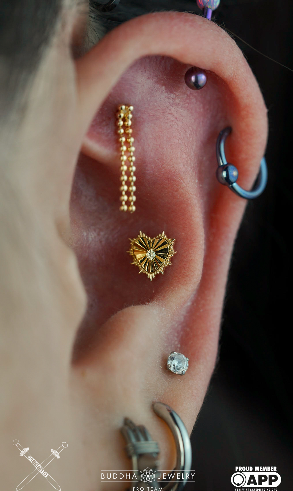Close-up of a person's ear with multiple earrings, including a gold chain, a heart-shaped earring, and a small gemstone, with a blurred background.