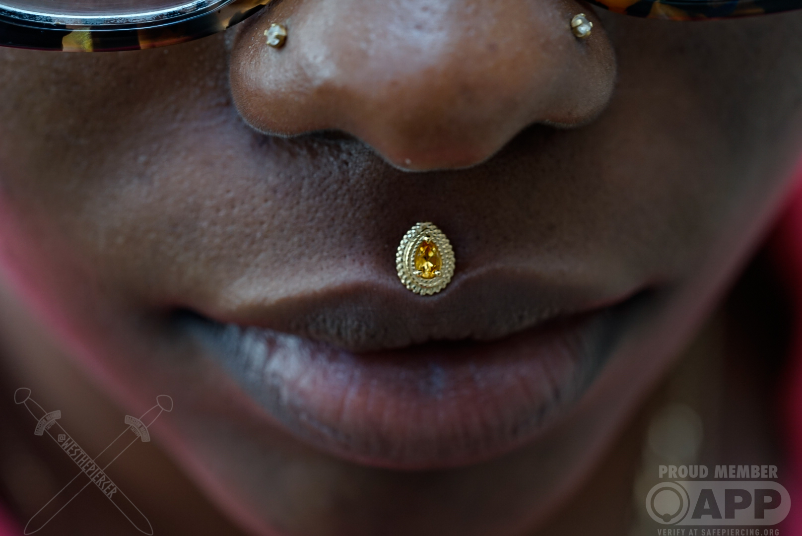 Close-up of a person's lips, nose, and a decorative nose ring with a yellow gemstone, with glasses partially visible.