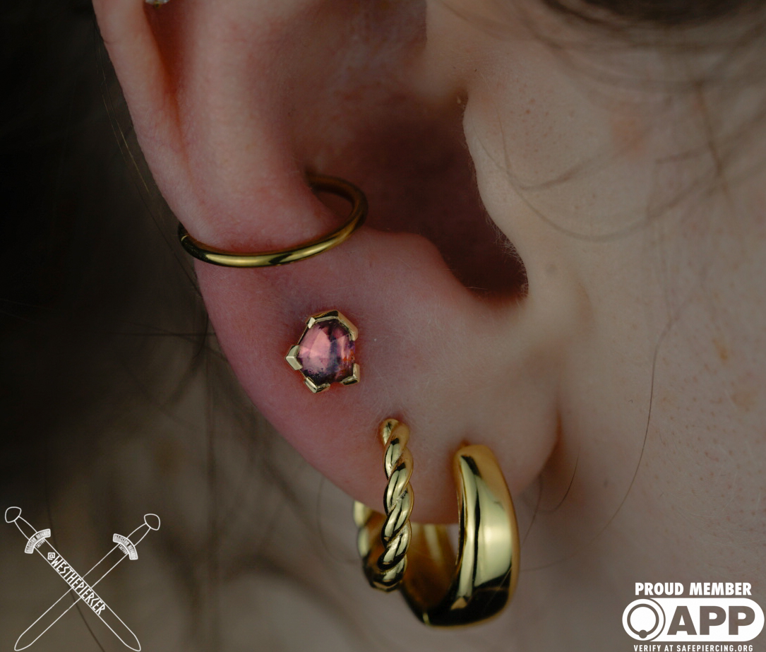 Close-up of a person's ear with multiple earrings, including a gold hoop, a pink gemstone stud, and a gold twisted hoop.