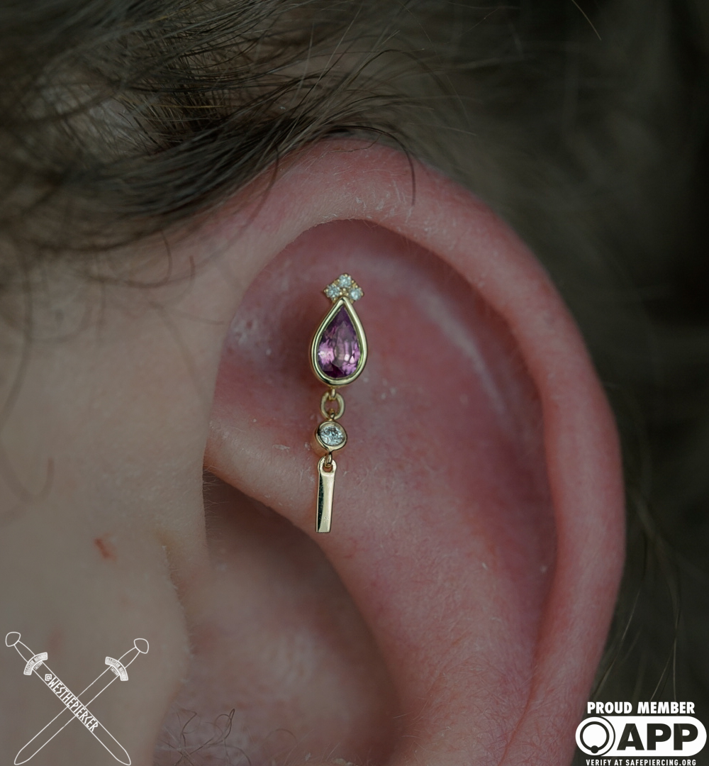 Close-up of a person's ear with a decorative earring featuring purple and clear gemstones, and a gold bar charm.