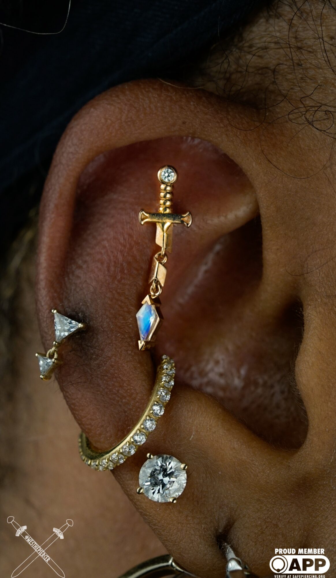 Close-up of a person's ear with multiple earrings, including a gold sword-shaped earring, a diamond stud, and a hoop with a large gemstone.
