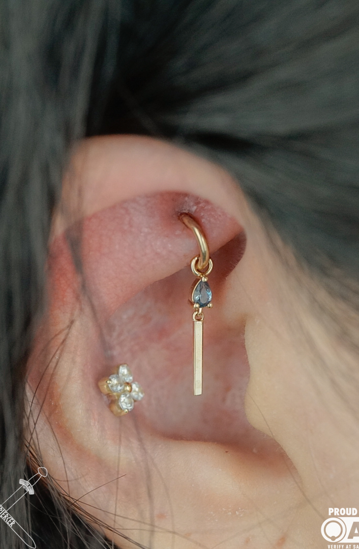Close-up of a person's ear with multiple earrings, including a gold hoop with a dangling blue gemstone and a flower-shaped cluster of small pearls or diamonds. Word count: 20