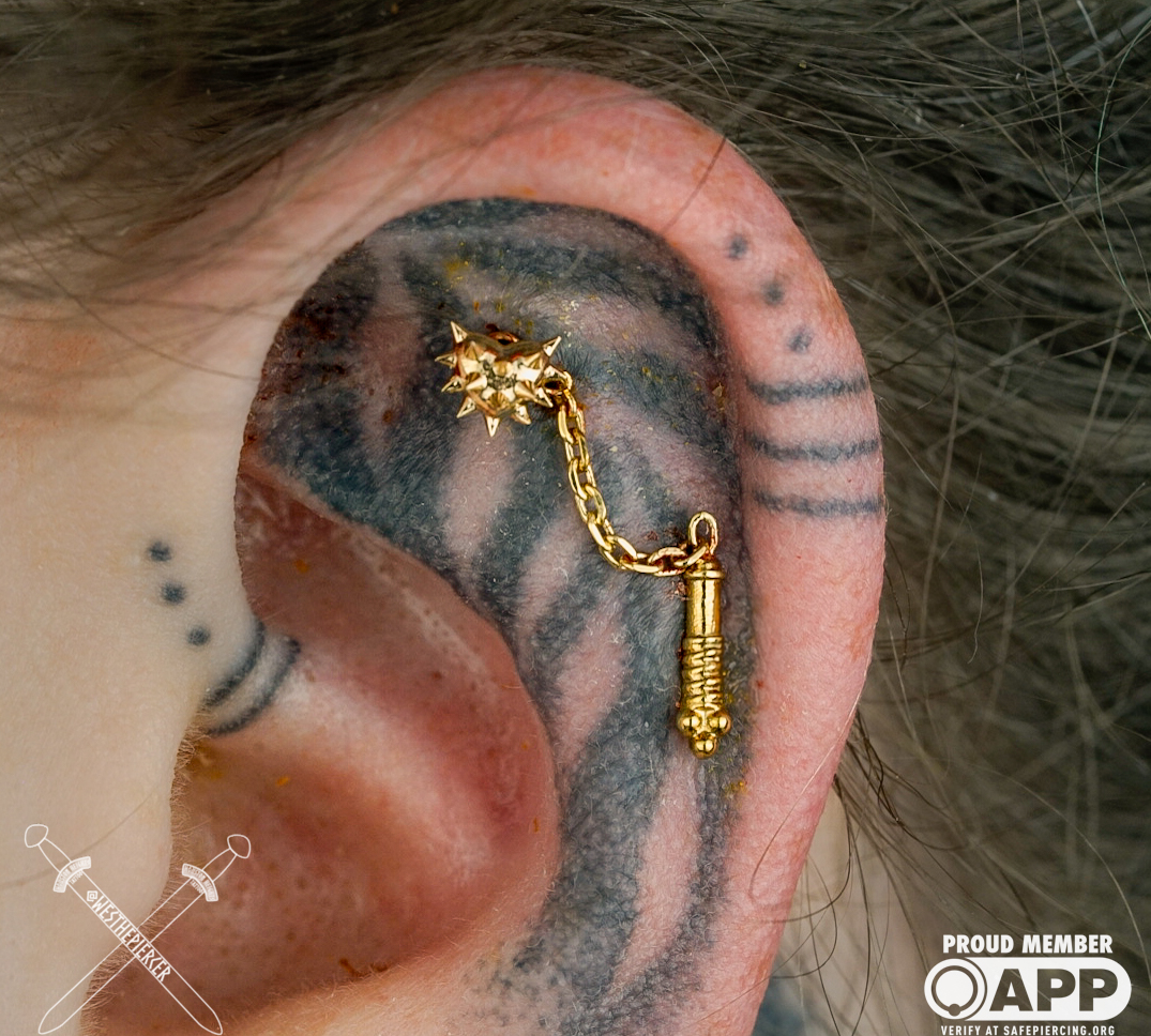 Close-up of a person's ear with a gold chain and pendant tattoo, and black ink tattoos on the ear and neck.
