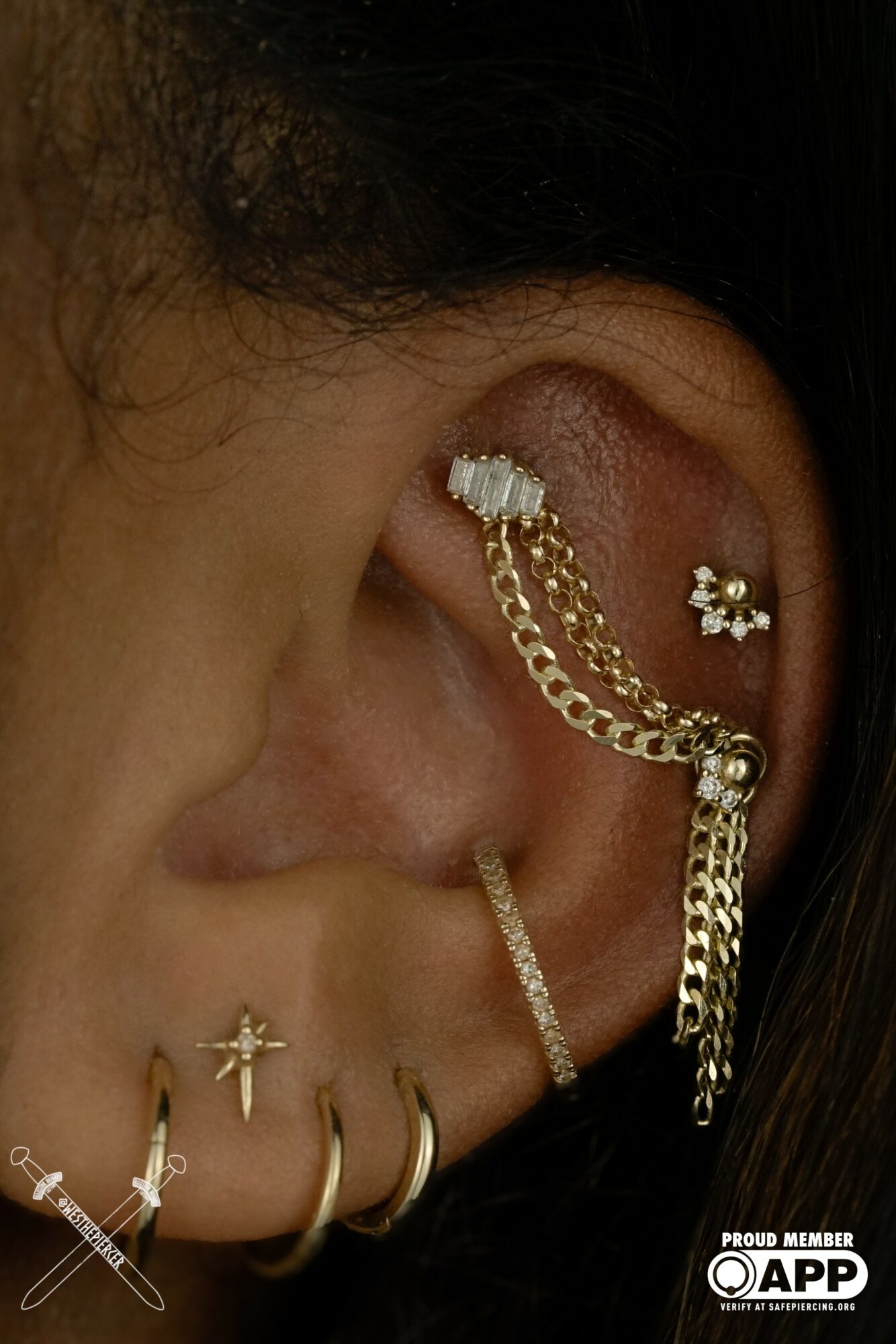 Close-up of a person's ear with multiple earrings, including chains and decorative studs, with visible hair and skin texture.