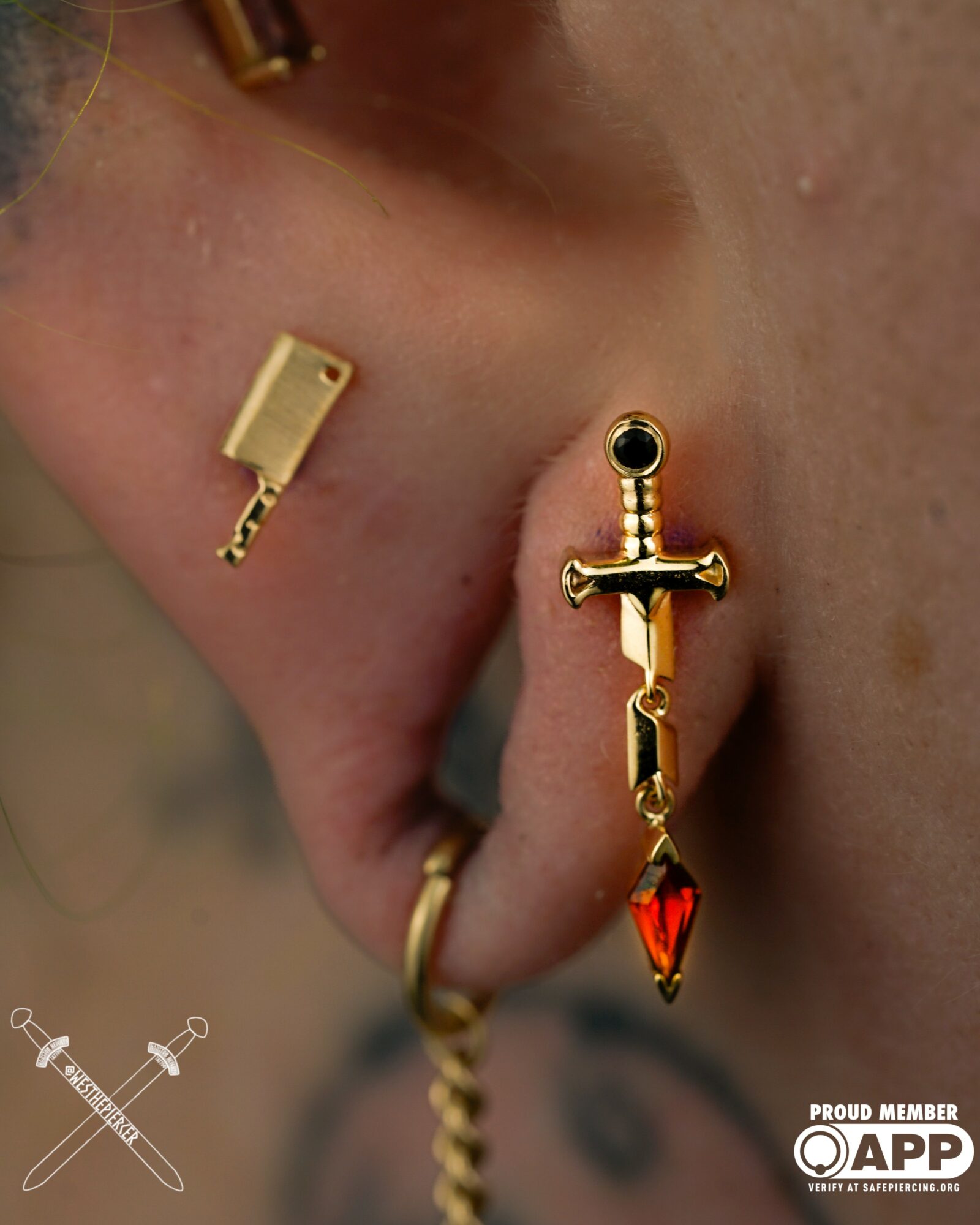 Close-up of a person's ear with multiple earrings, including a gold sword-shaped earring with a red gemstone, and a gold chain earring. Word count: 20