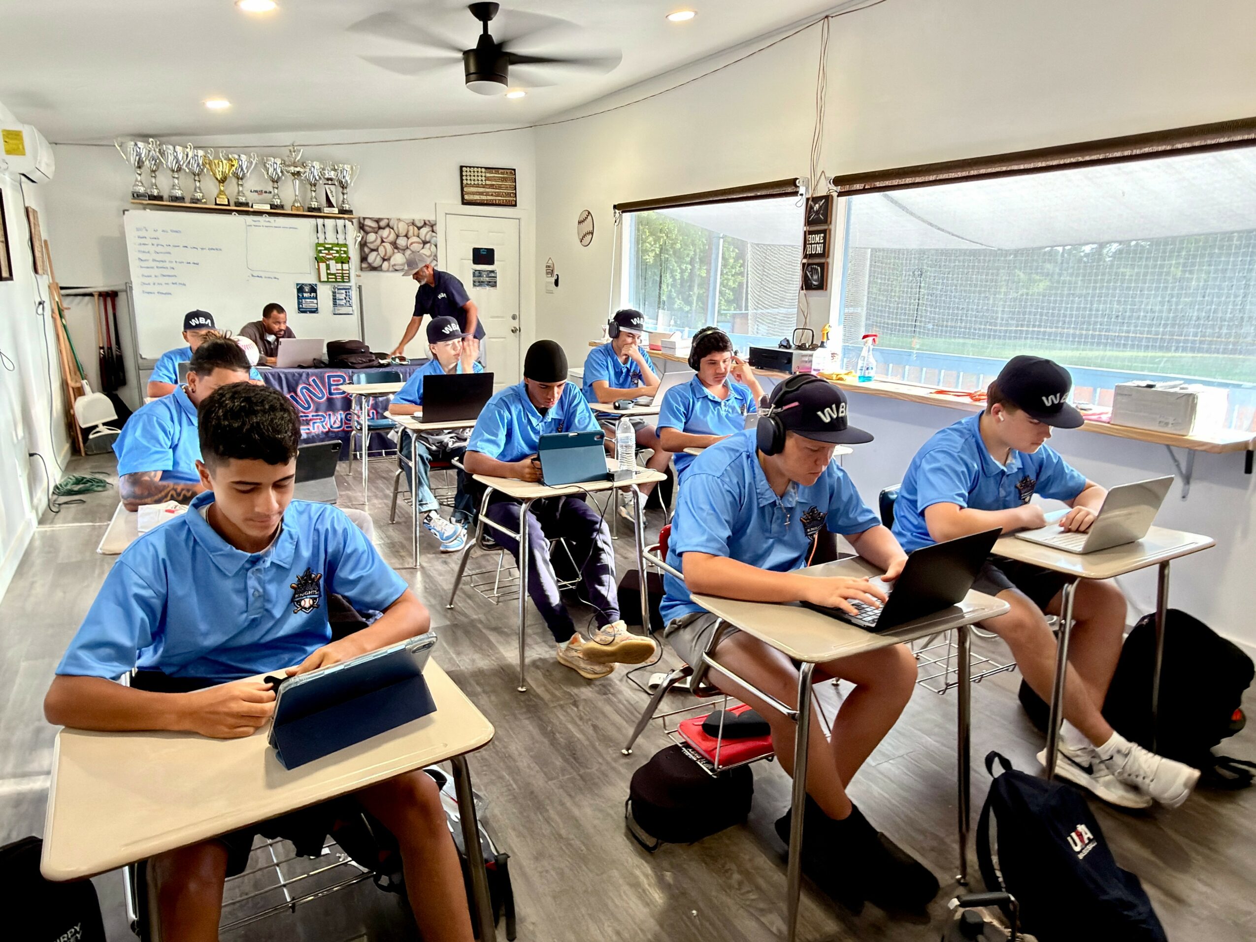Classroom with students sitting at desks, wearing blue uniforms, working on tablets, with a teacher at the whiteboard.