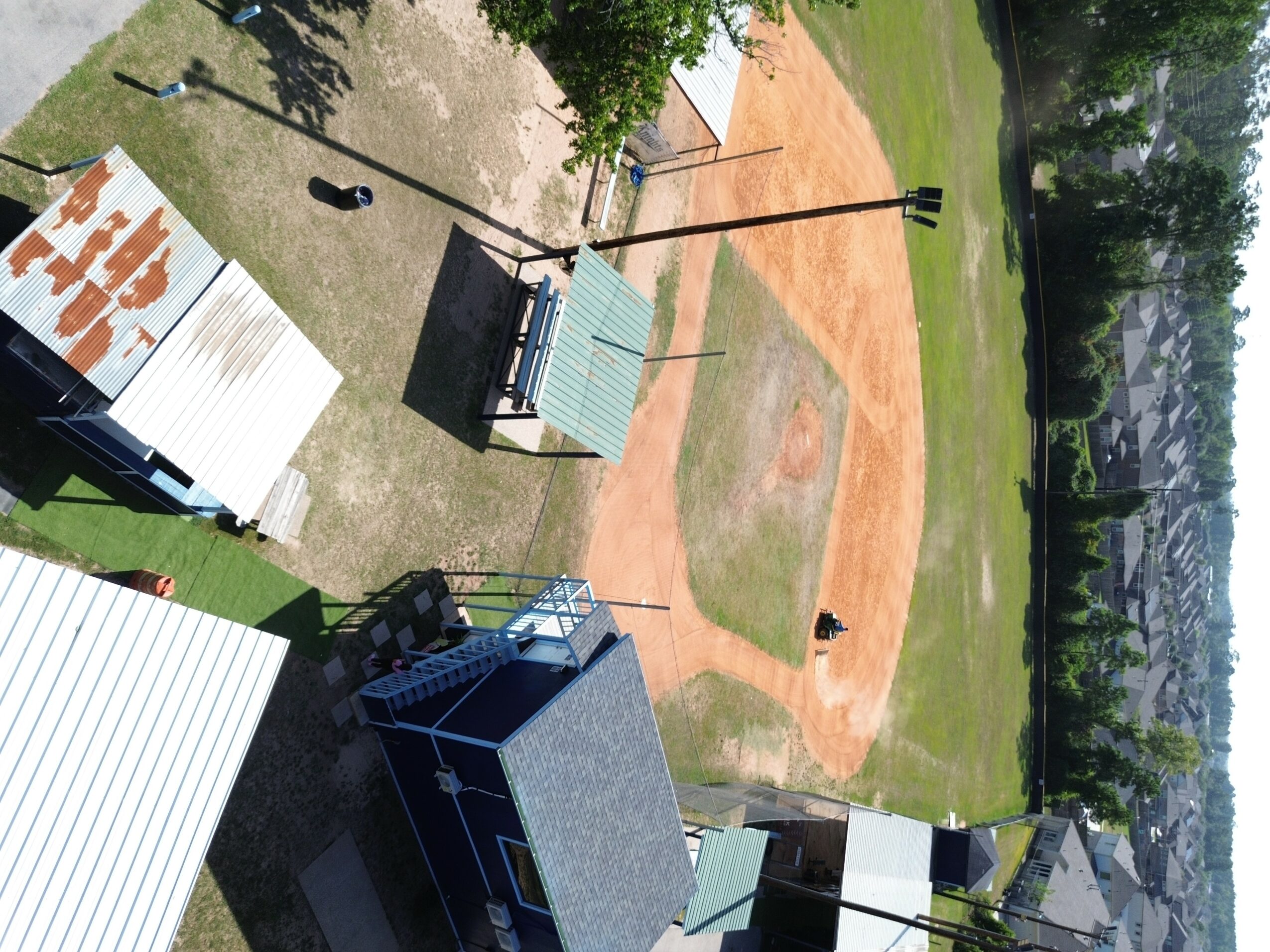 Baseball field with dirt infield, grass outfield, and surrounding trees, viewed from above.