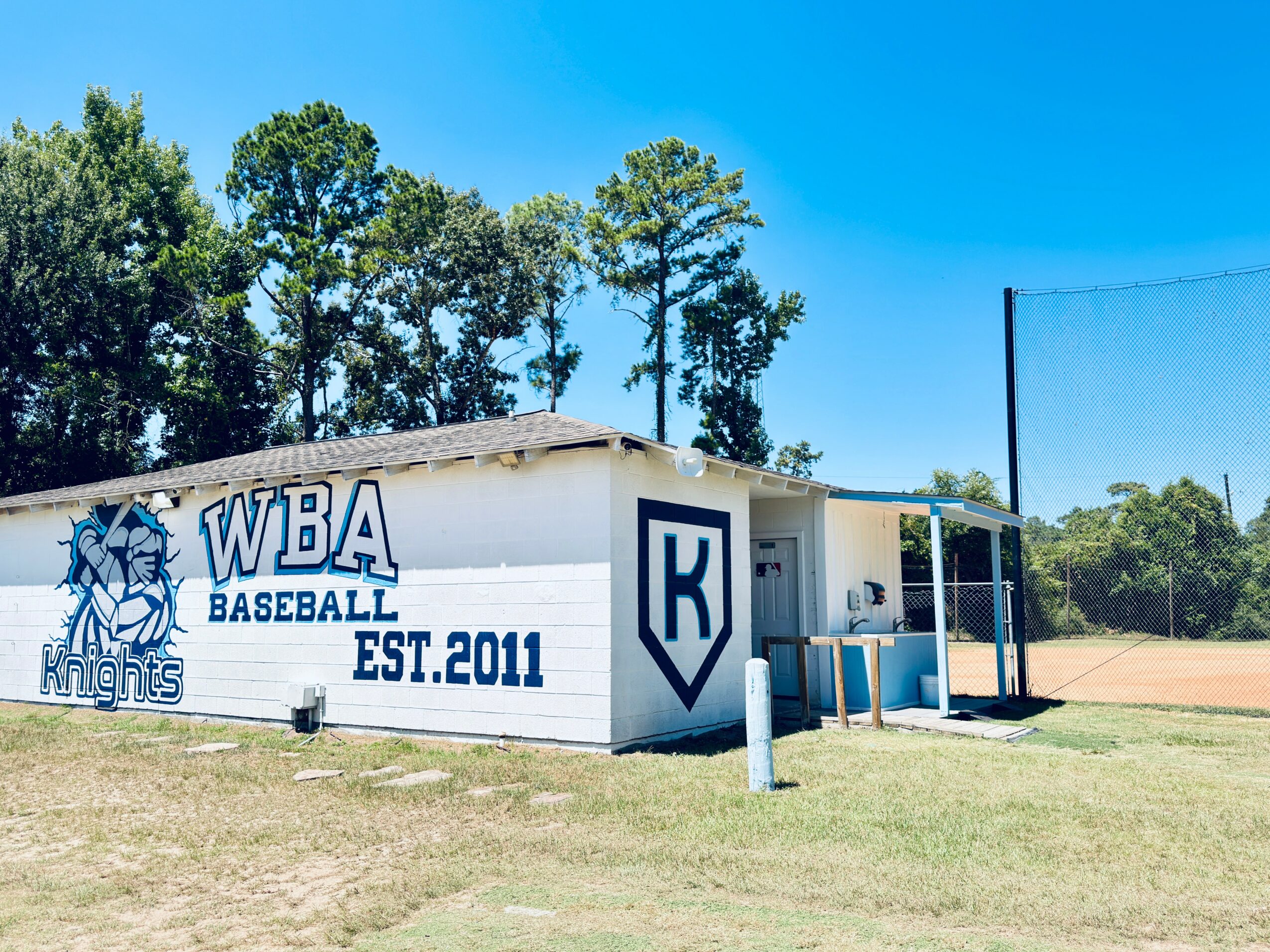 Baseball field building with WBA Baseball Knights logo, trees in background, blue sky, and a small covered area.