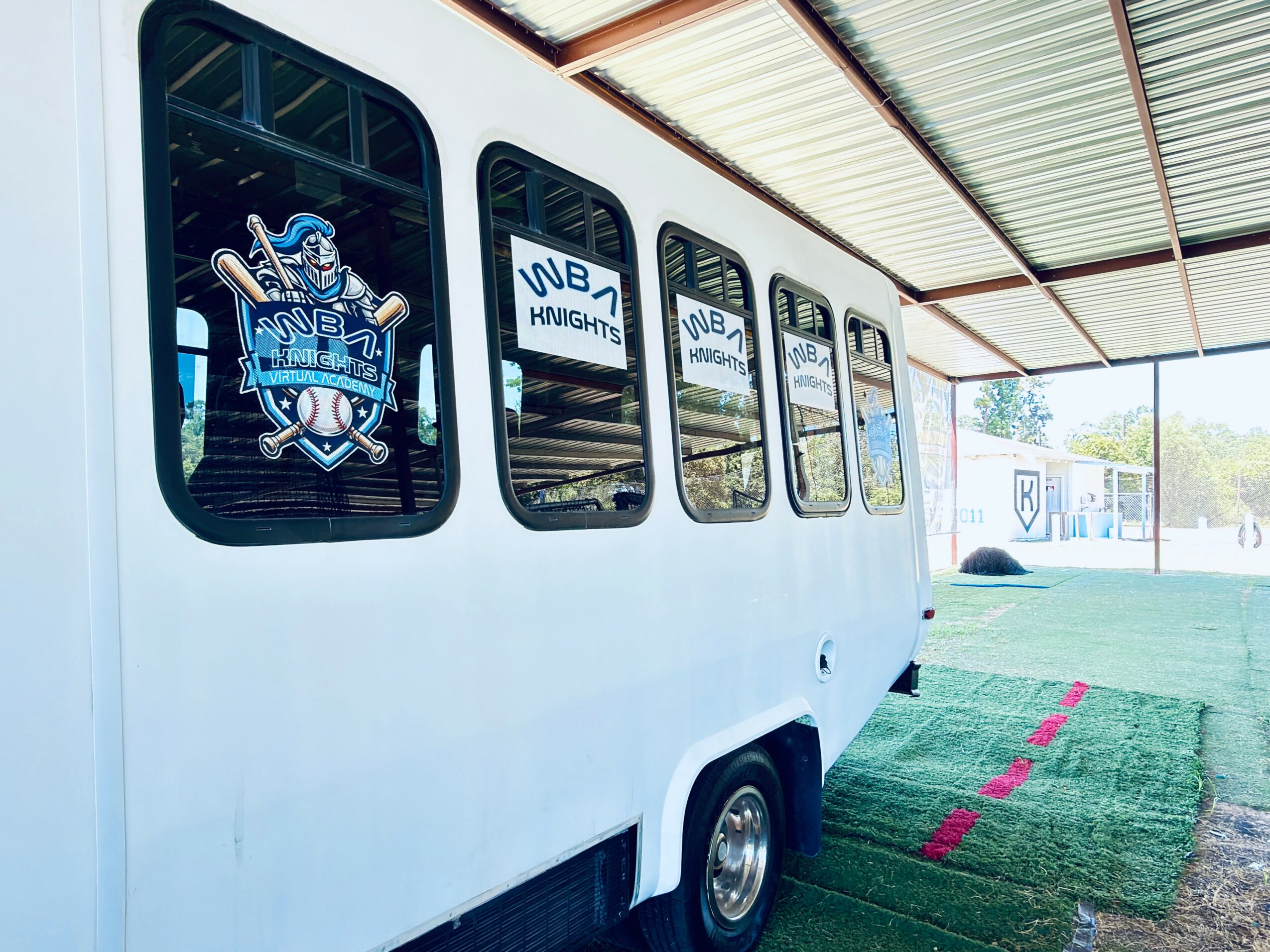 Side view of a white bus with windows, parked under a metal roof, with a grassy area nearby.