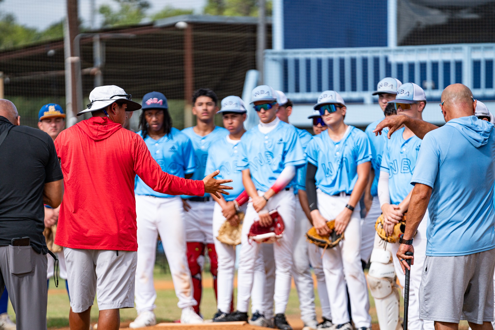 Group of young baseball players in blue uniforms listening to coach in red jacket outdoors.