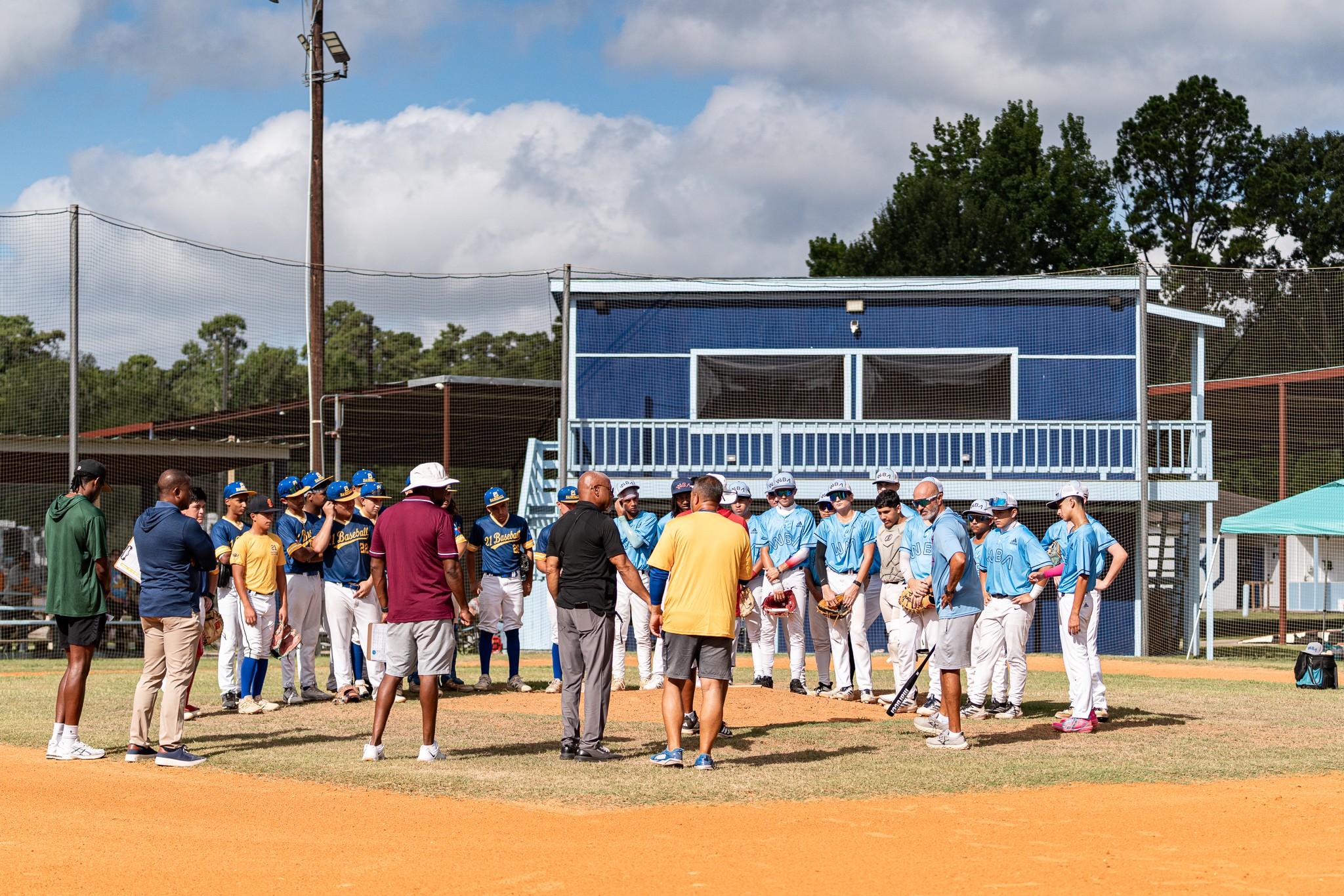 Group of people gathered on a baseball field near a blue building with a balcony, under a partly cloudy sky.