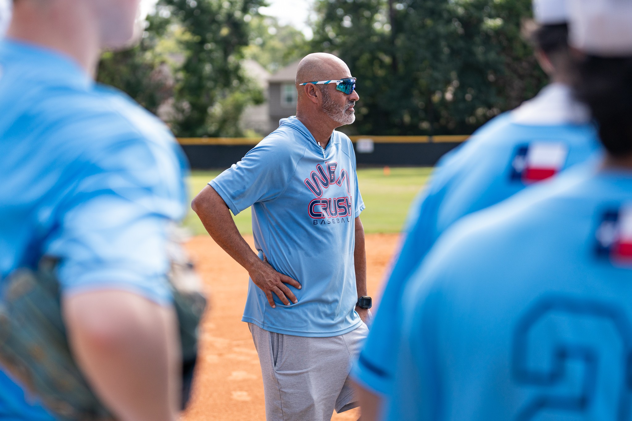 Bald man in sunglasses and light blue shirt stands on a baseball field with hands on hips, surrounded by players in blue uniforms.