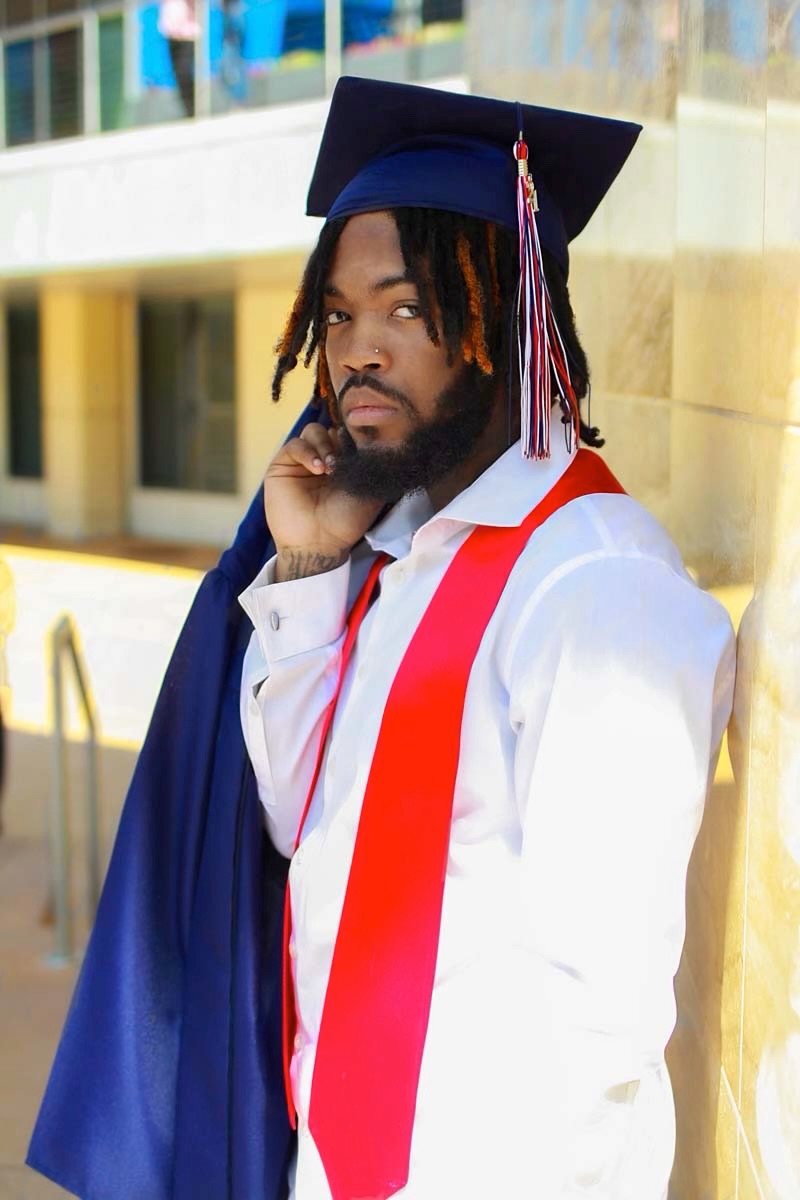 Young man in graduation cap and gown, standing outdoors, looking at camera, with hand near face, sunlight, building background.