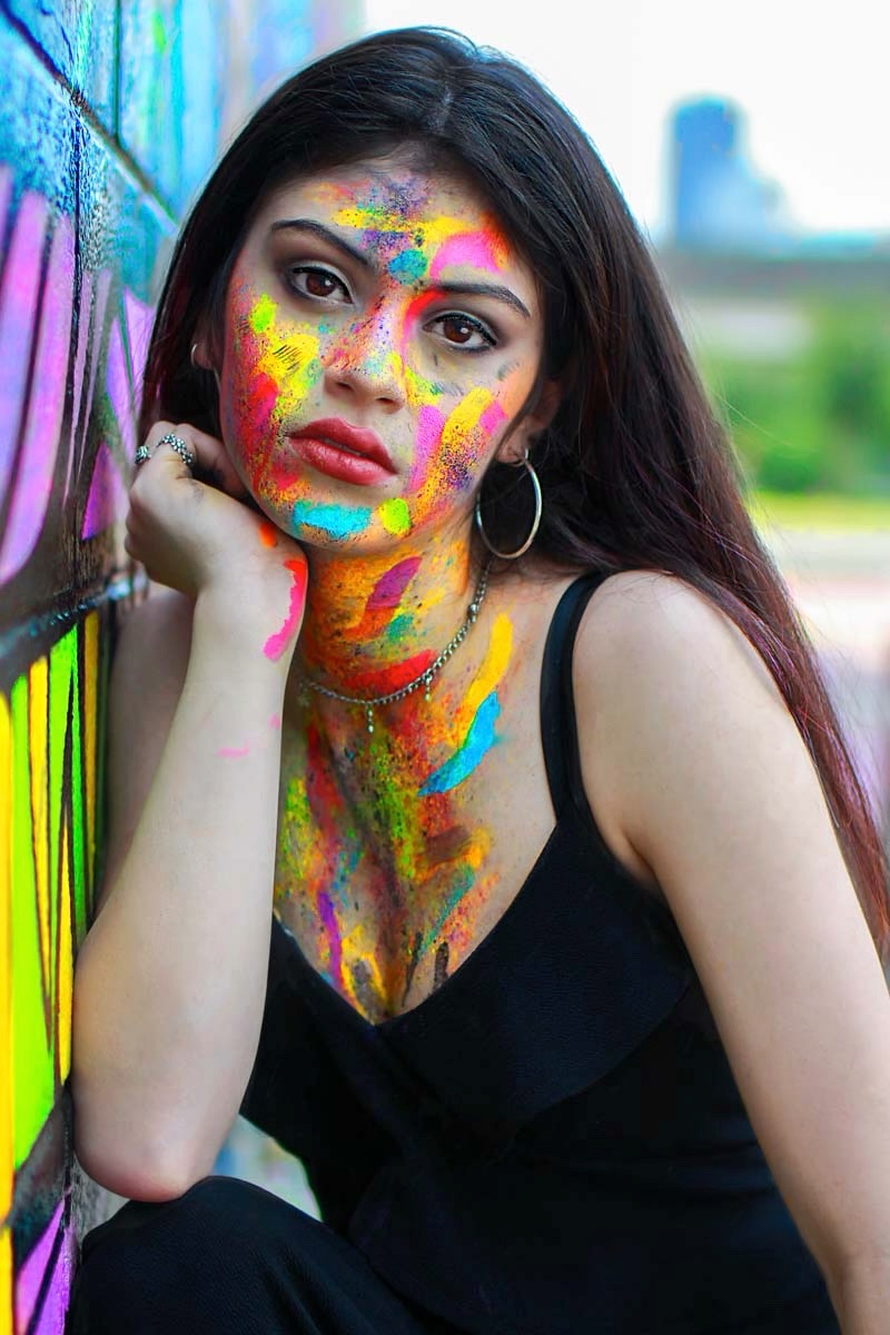 Young woman with colorful paint on face and neck, resting her chin on her hand, outdoors with blurred background.