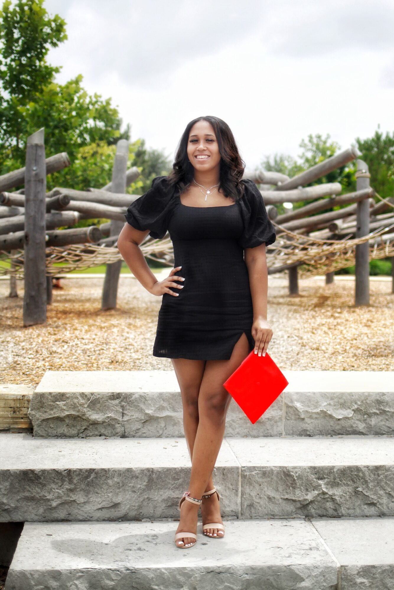 Woman in black dress holding a red clutch, standing outdoors on steps with wooden fence and trees behind her.