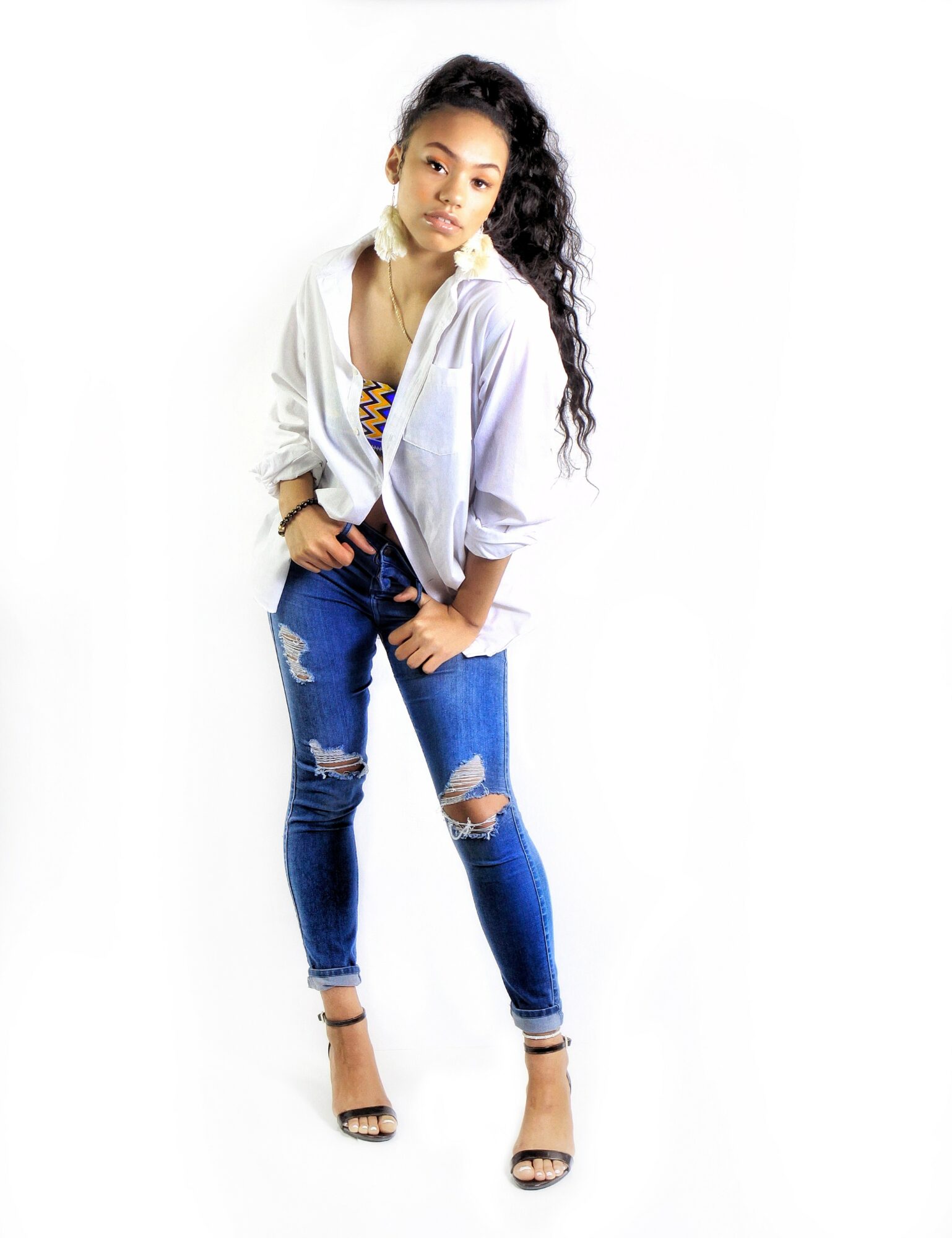 Young woman with long curly hair wearing a white jacket, striped top, ripped jeans, and high heels, standing against a white background.