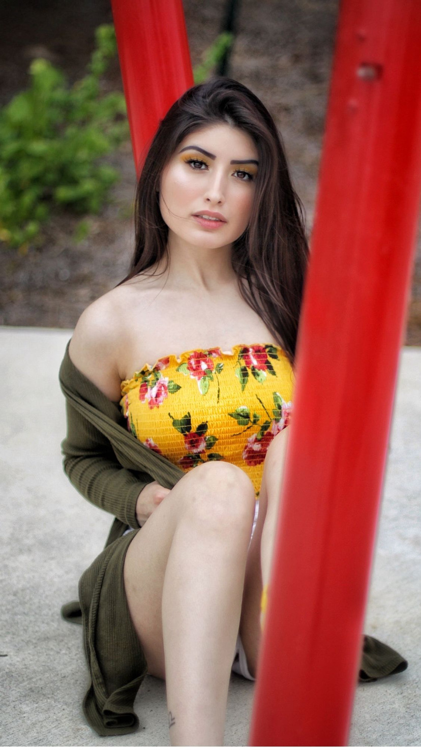 Young woman with long dark hair sitting outdoors, wearing a yellow floral strapless top and an open jacket, with red playground equipment nearby.
