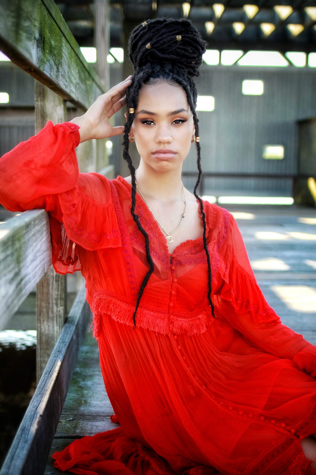 Young woman with braided hair in a red dress leaning against a wooden railing outdoors.