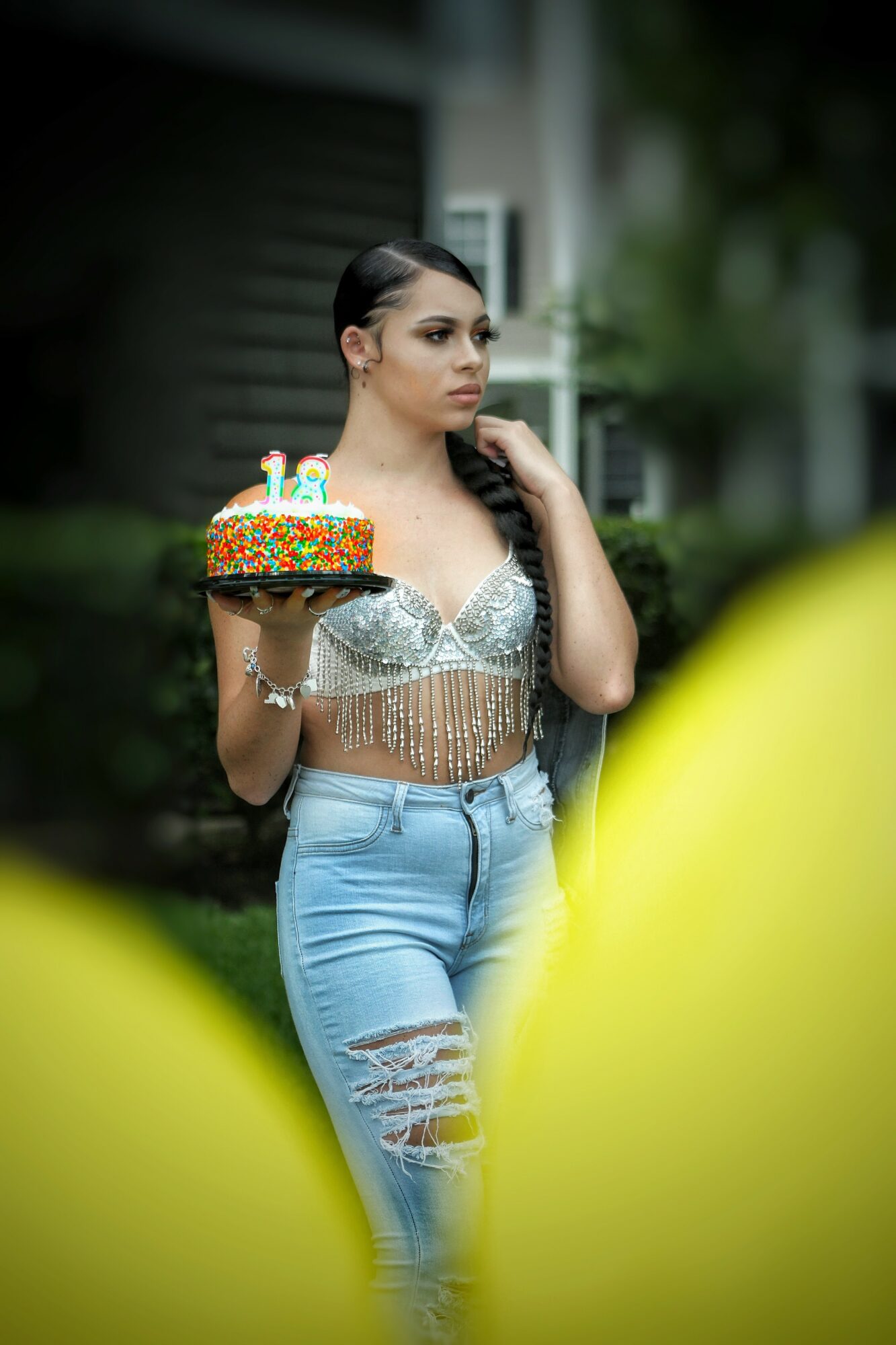 Woman holding a birthday cake with lit candles outdoors, wearing a silver top and ripped jeans, with blurred background.