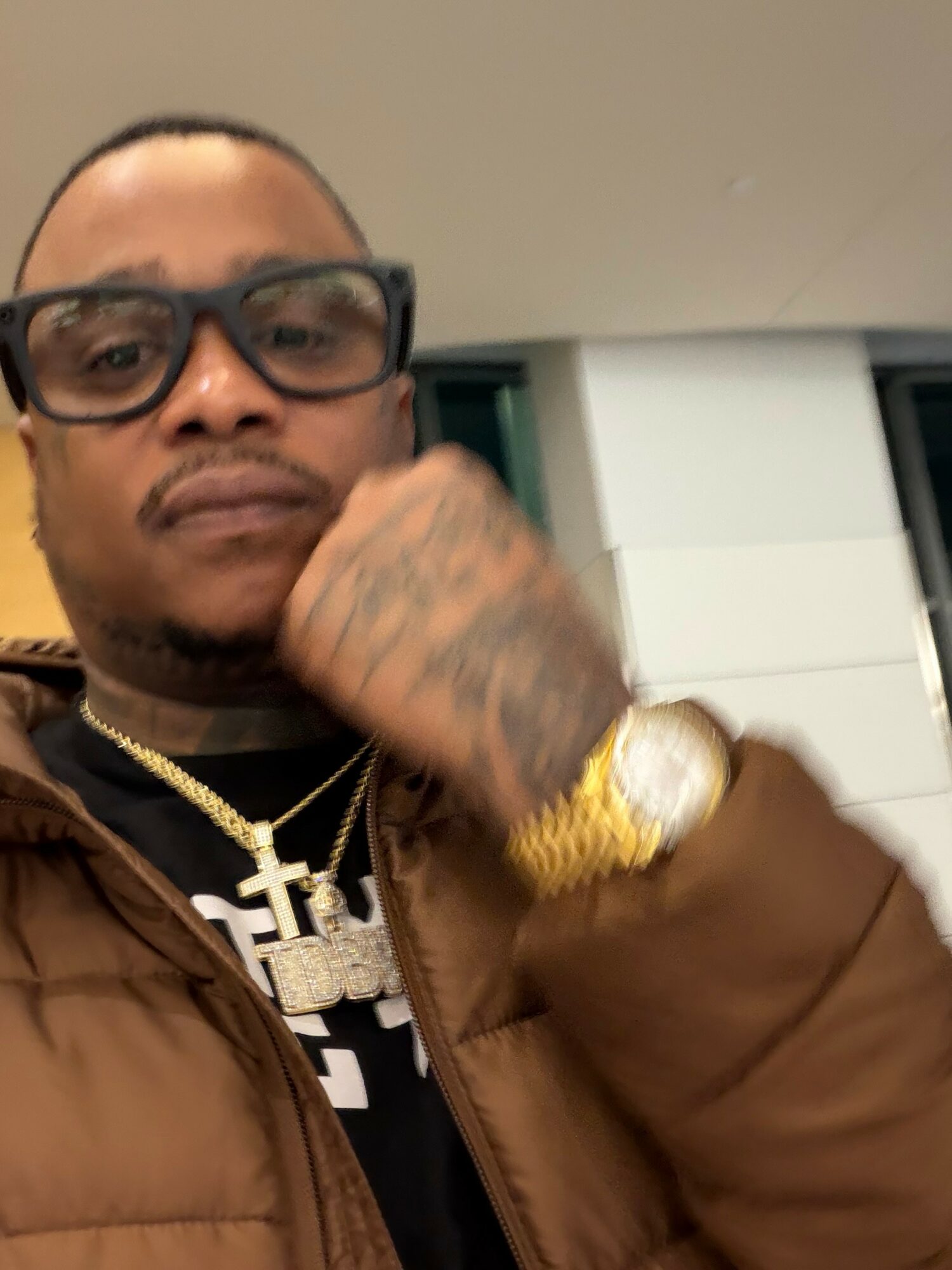 Person with glasses, gold watch, and chain, resting chin on hand, in an indoor setting.
