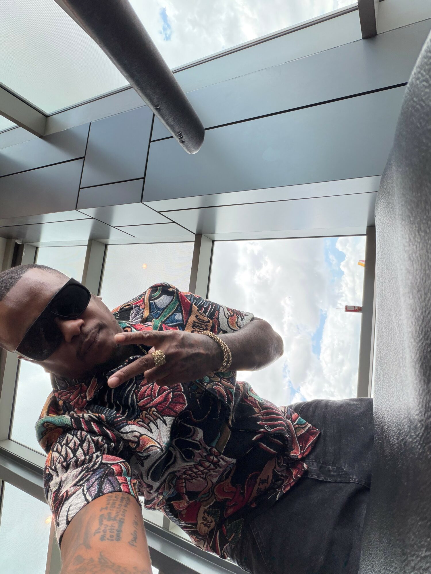 Person lying on the floor wearing sunglasses and a patterned shirt, with a view of the sky through a window.