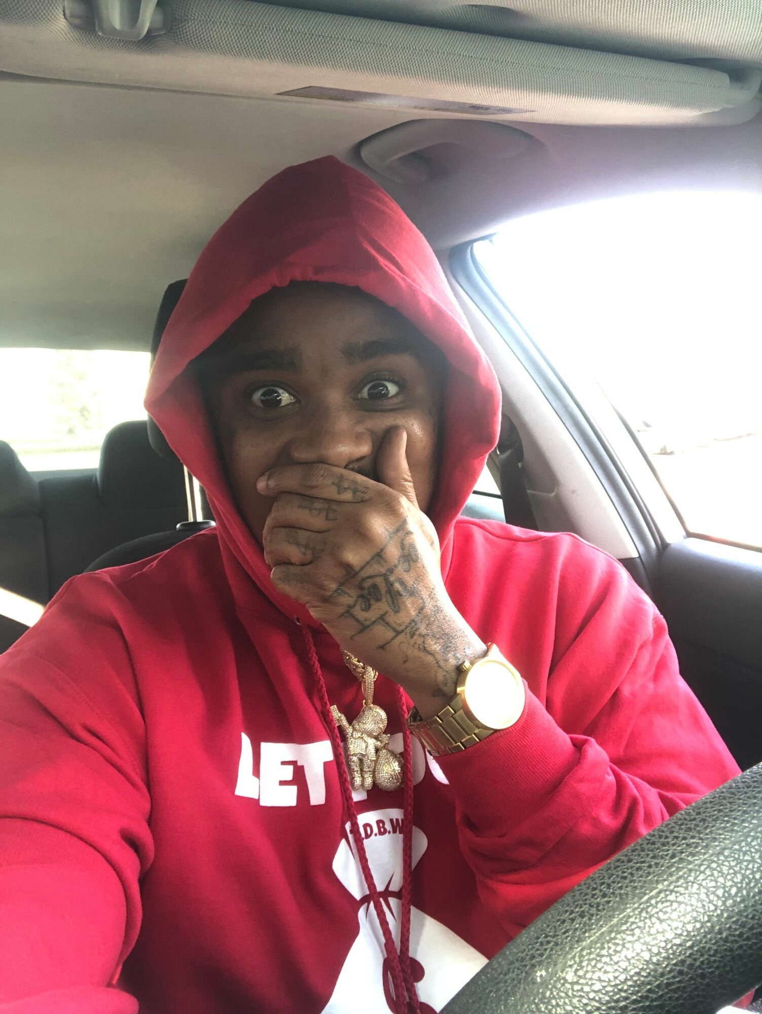 Person wearing a red hoodie with a hood over head, sitting in a car, covering mouth with hand, tattoos visible on hand, watch on wrist.