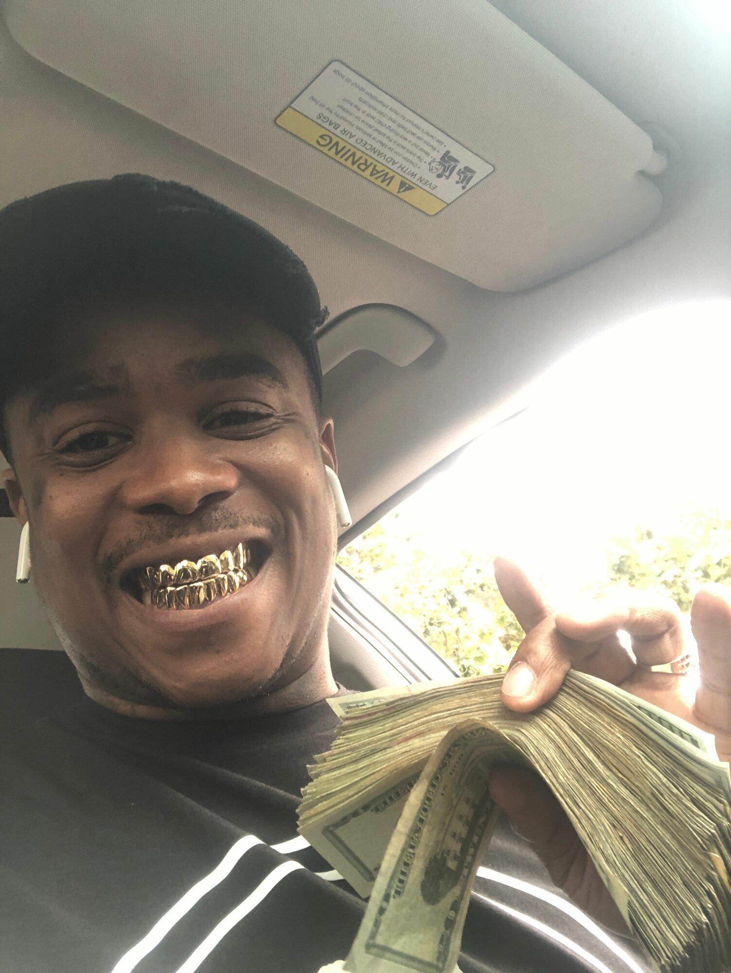 Person smiling with gold teeth, holding a large stack of money inside a car, wearing a striped shirt.
