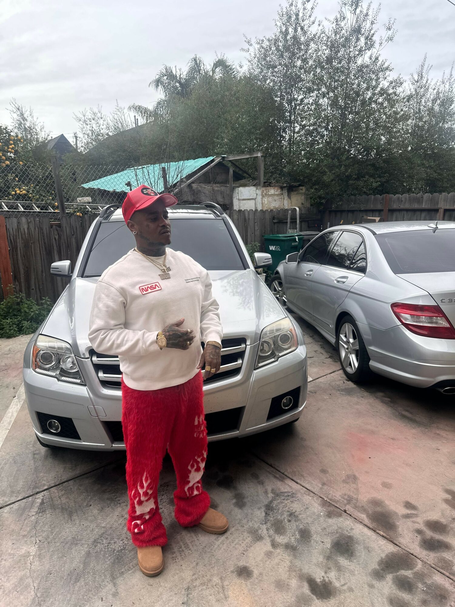 Person in red pants and beige jacket standing next to a silver car, holding a phone, in a driveway with other cars and trees.