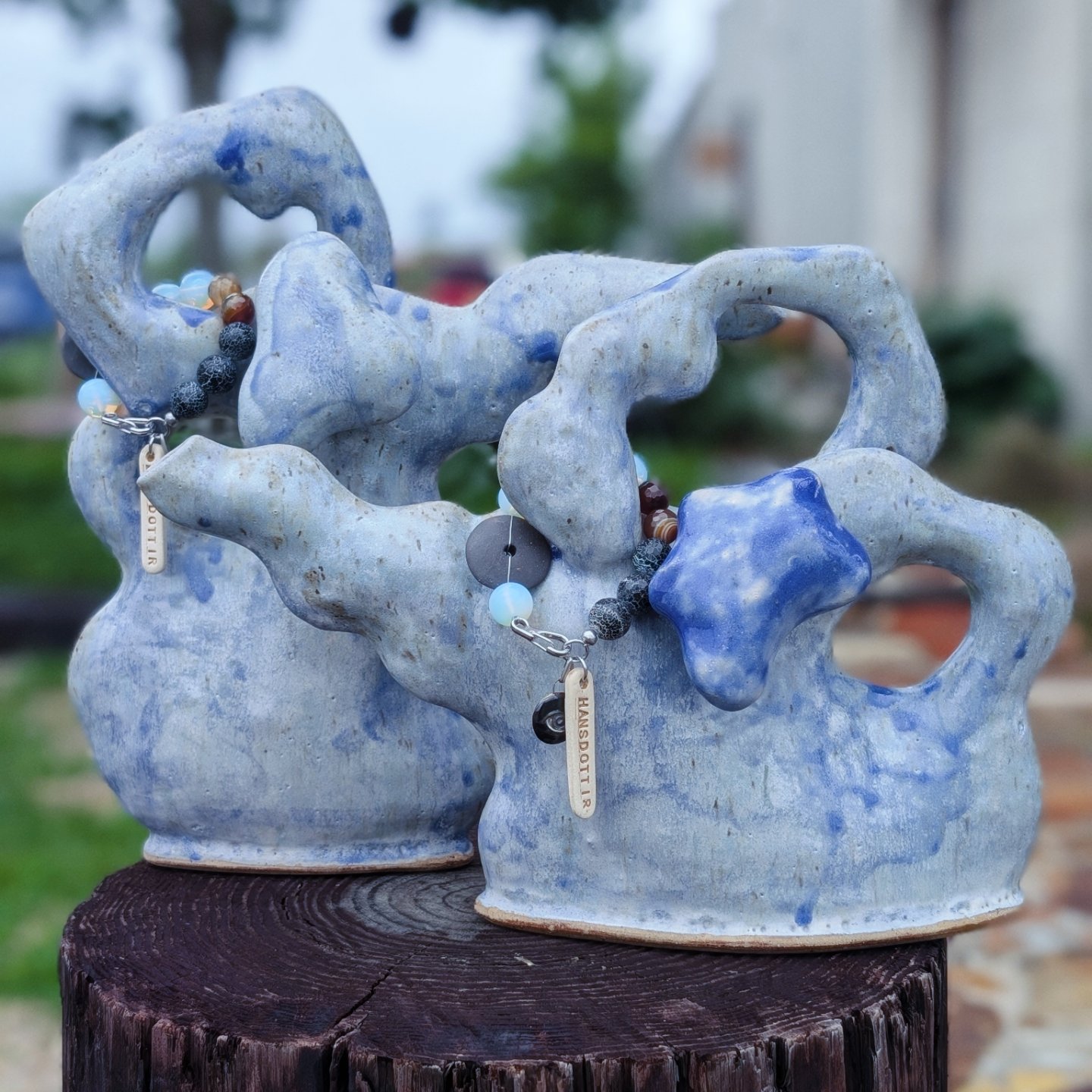 Three blue and white ceramic sculptures resembling abstract trees or coral, with necklaces hanging on them, placed outdoors on a wooden surface.