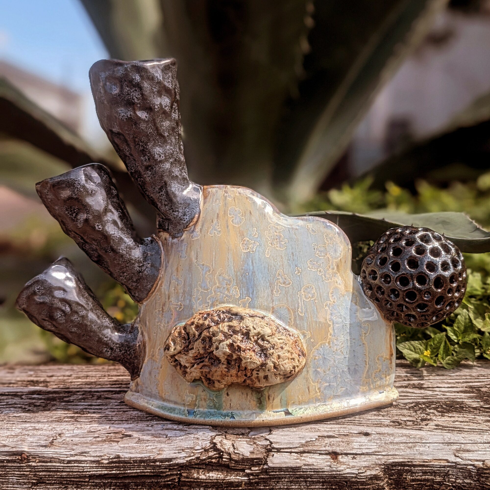 Decorative ceramic sculpture with dark textured elements, a round black object, and a rough brown feature, placed outdoors on a wooden surface.